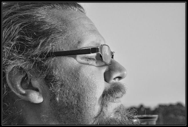 Close-up side profile of a man with glasses, a beard, and long hair, looking to the right in black and white.