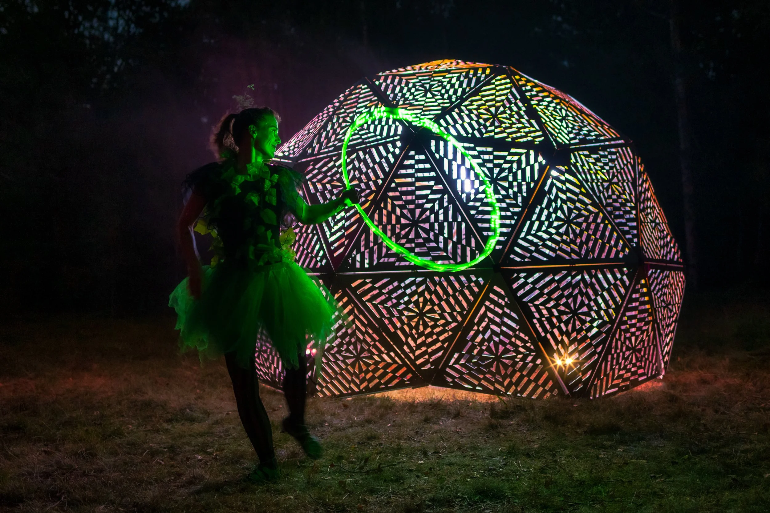 A woman in a tutu and puffy shirt performs with a green glow stick in front of a large illuminated geometric sphere at night.