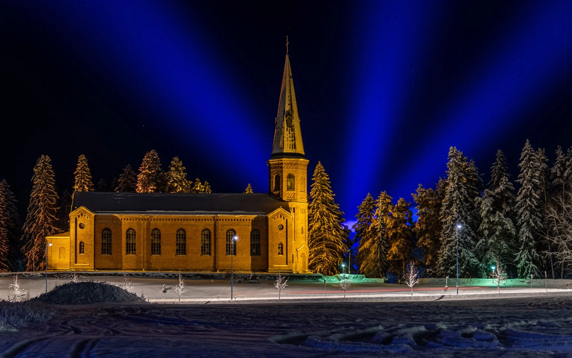 Night view of a lit church with a tall steeple, surrounded by snow-covered trees and a snow-covered field, under a dark sky with blue light beams.