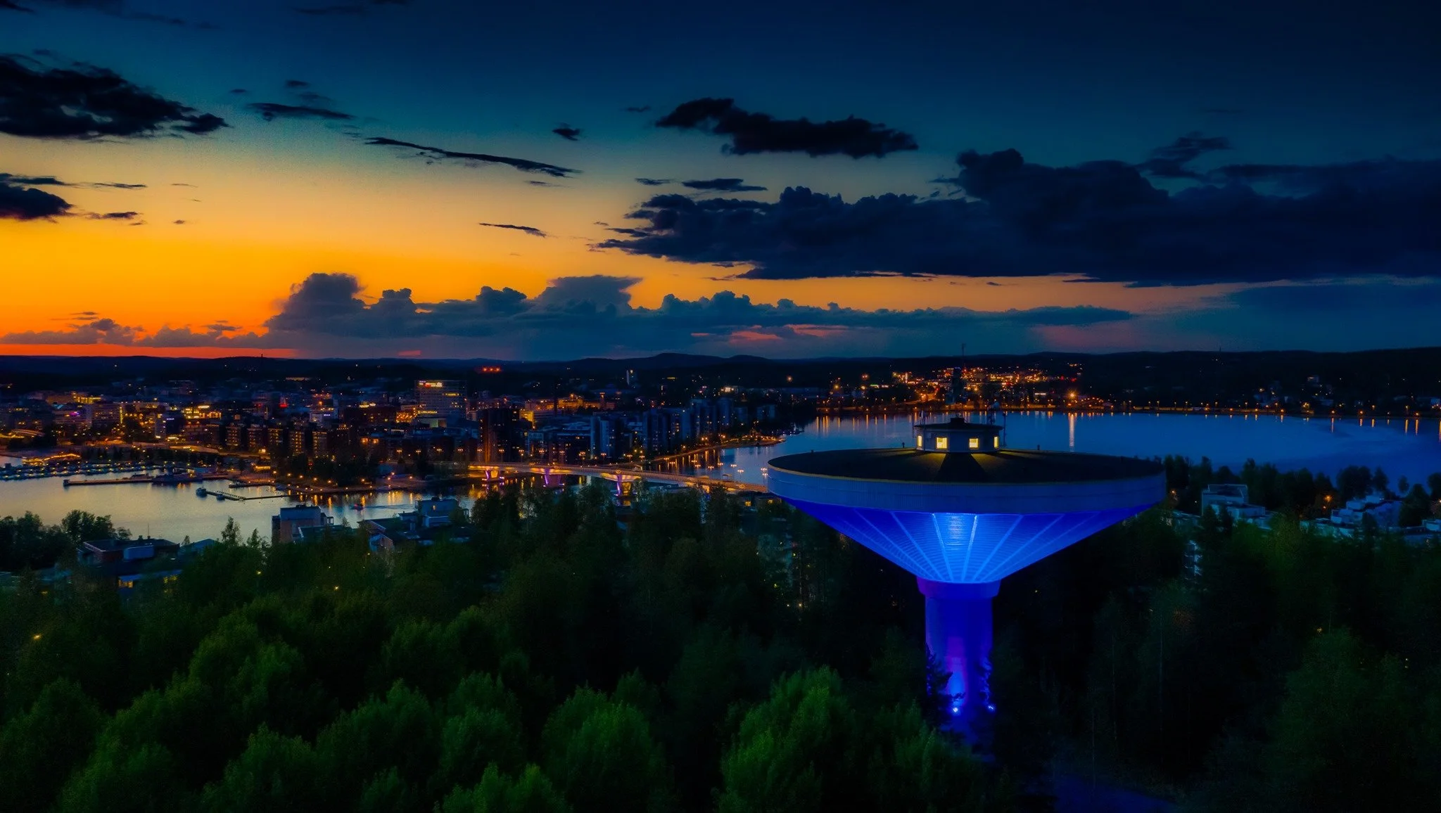A city skyline at dusk with a body of water, a uniquely shaped observation tower illuminated in blue, and a colorful sunset with clouds in the sky.
