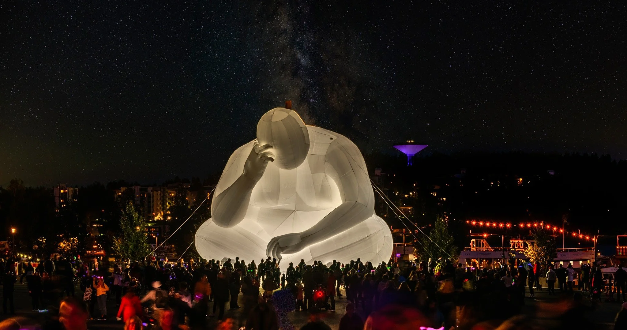 A large illuminated sculpture of a human figure sitting and holding an egg to their ear, displayed outdoors at night with a starry sky in the background and a crowd of people gathered around.