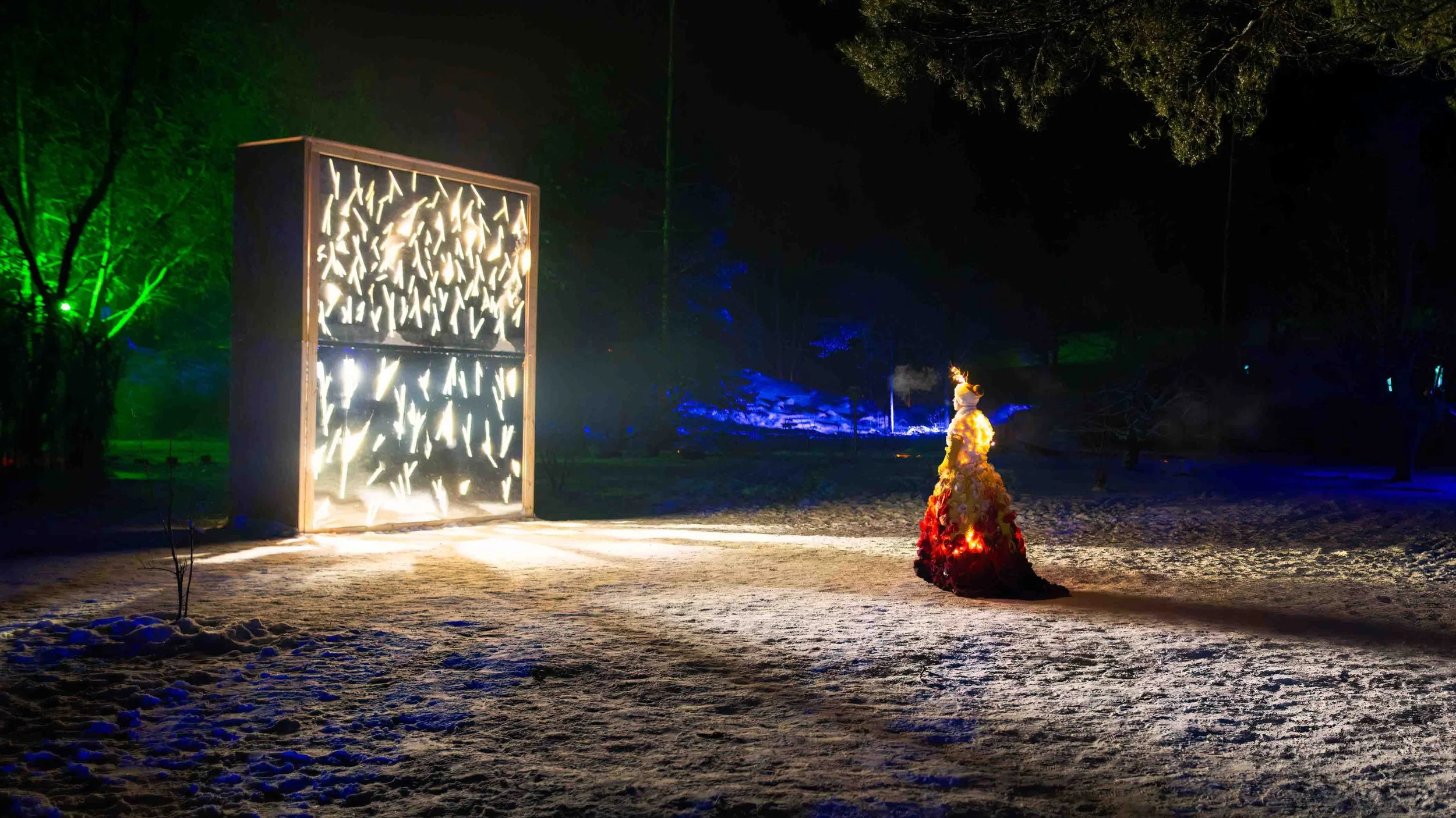 Outdoor nighttime scene with illuminated sculpture resembling a dress made of flowers, and a large artistic light installation with abstract shapes. Snow covers the ground, with trees illuminated in different colors in the background.