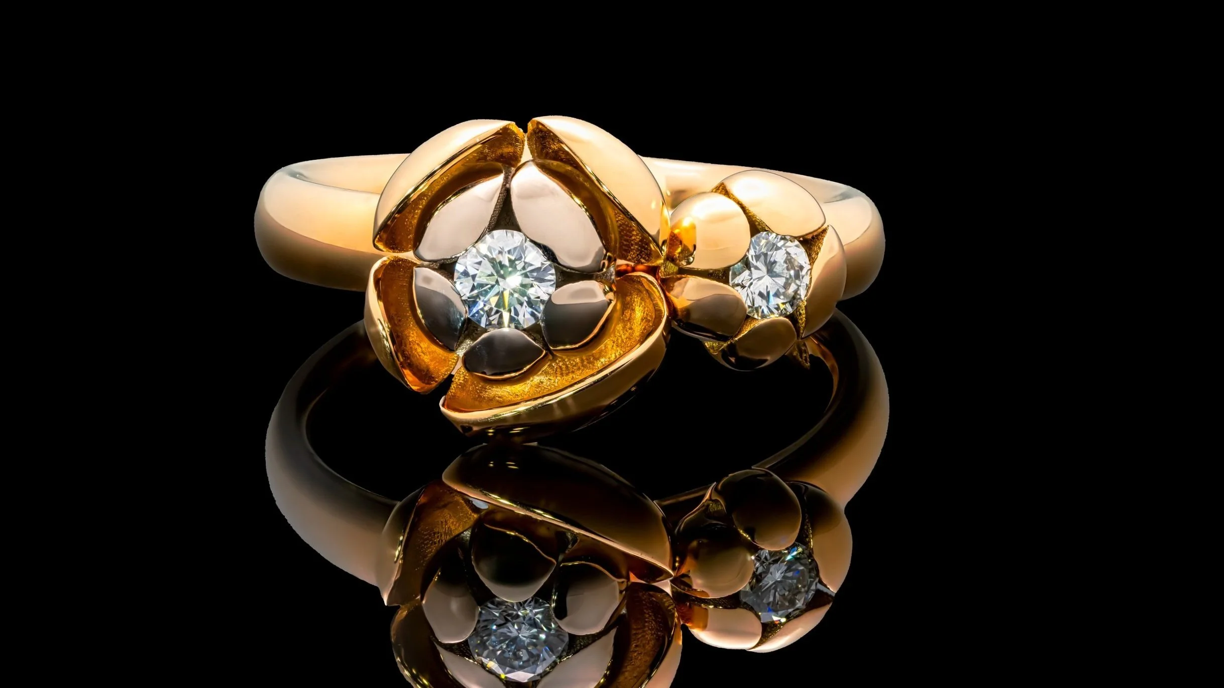 A gold ring with a floral design featuring two large diamonds, set against a black background with a reflection below.
