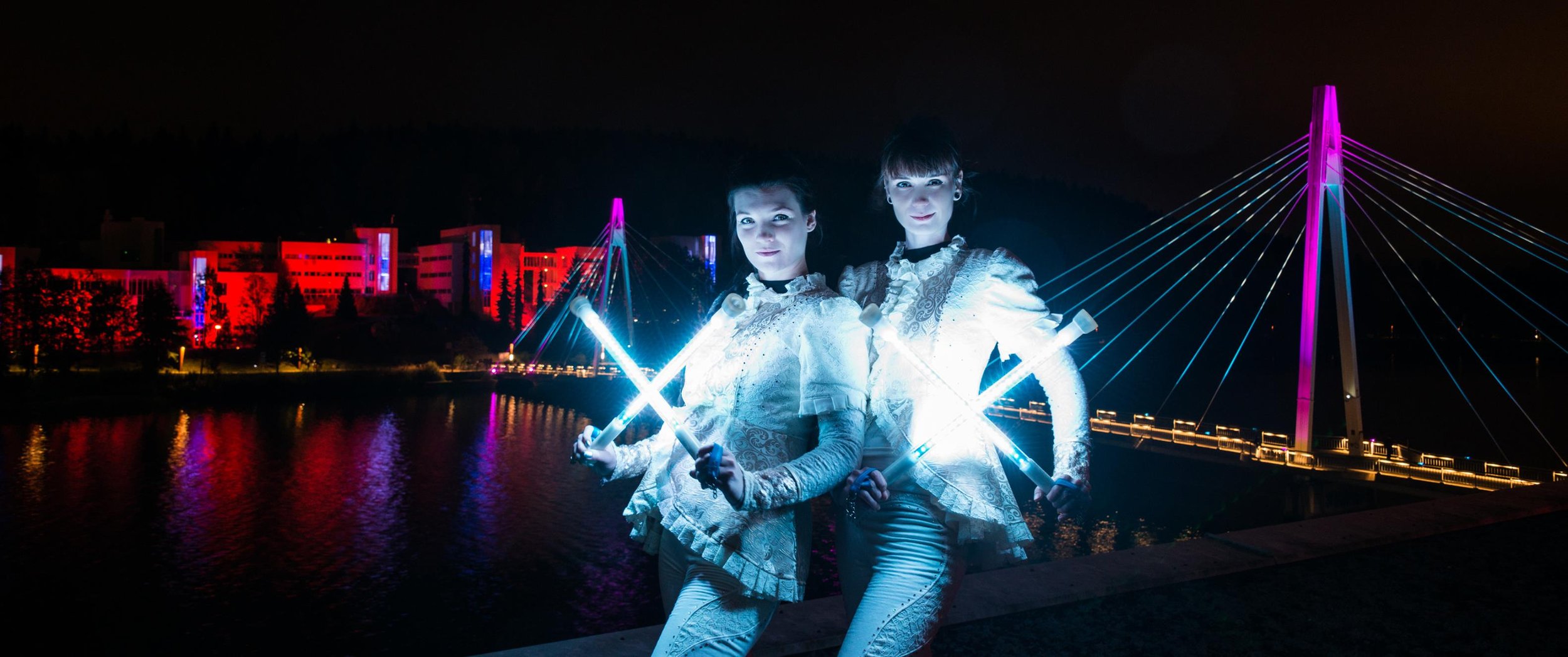 Two women with illuminated batons standing at night near a river, with a brightly lit modern bridge and colorful city buildings with red and purple lights in the background.