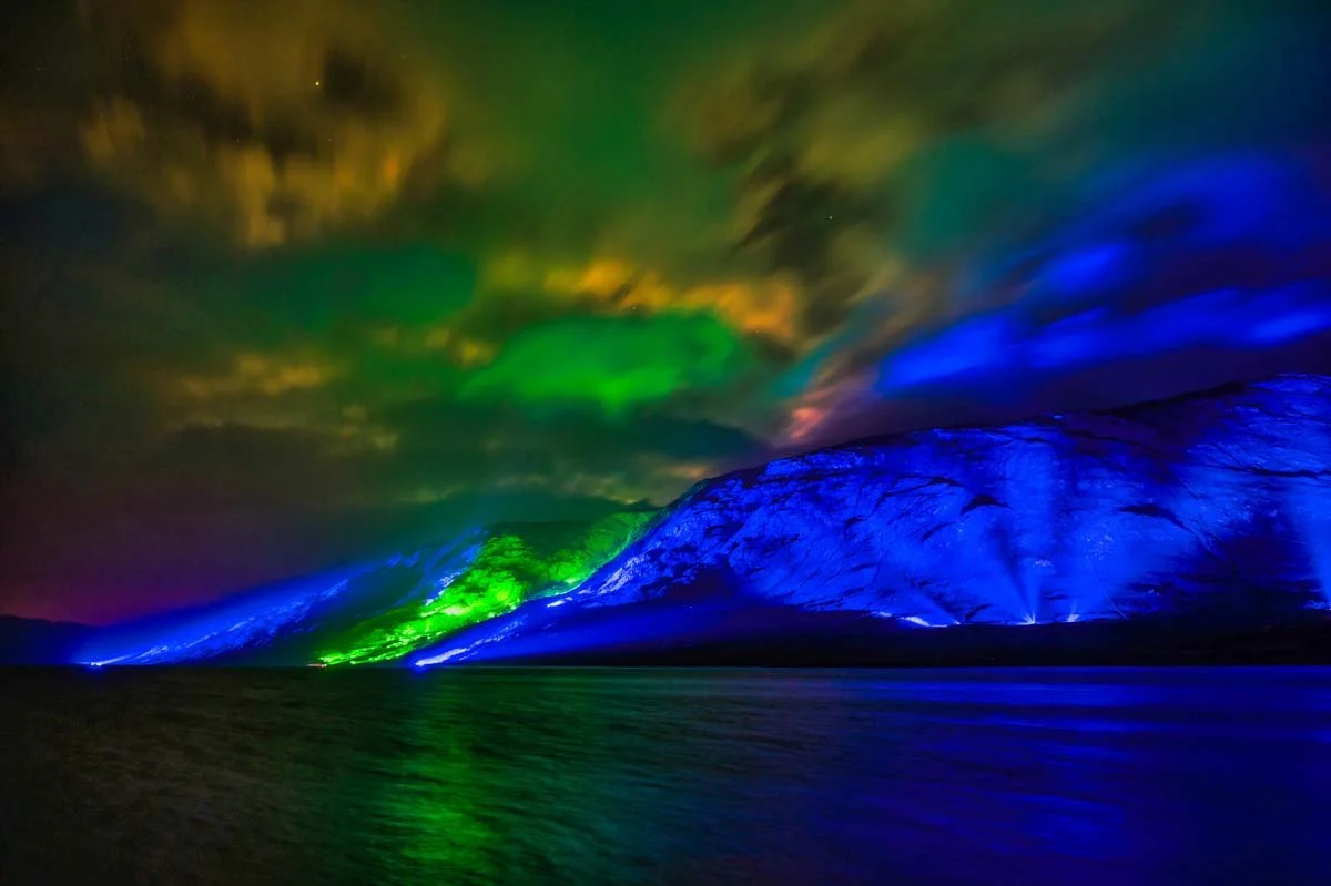Nighttime scene of a mountain reflecting colorful lights in the water, with the mountain illuminated in blue, and the sky shows green and orange auroras or light displays.