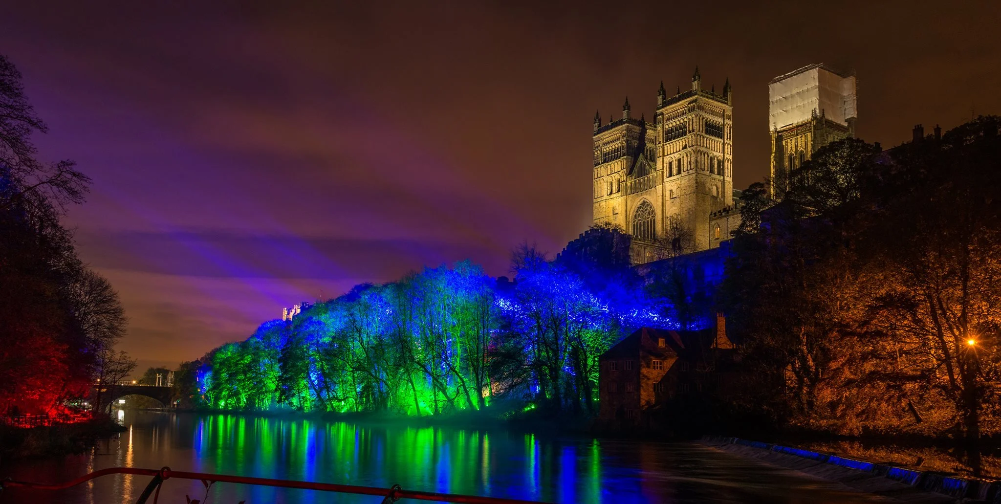 Night view of a historic castle illuminated with yellow lights, situated on a hill above a river. Trees along the riverbank are lit with blue, green, and red lights, creating a colorful reflection in the water.