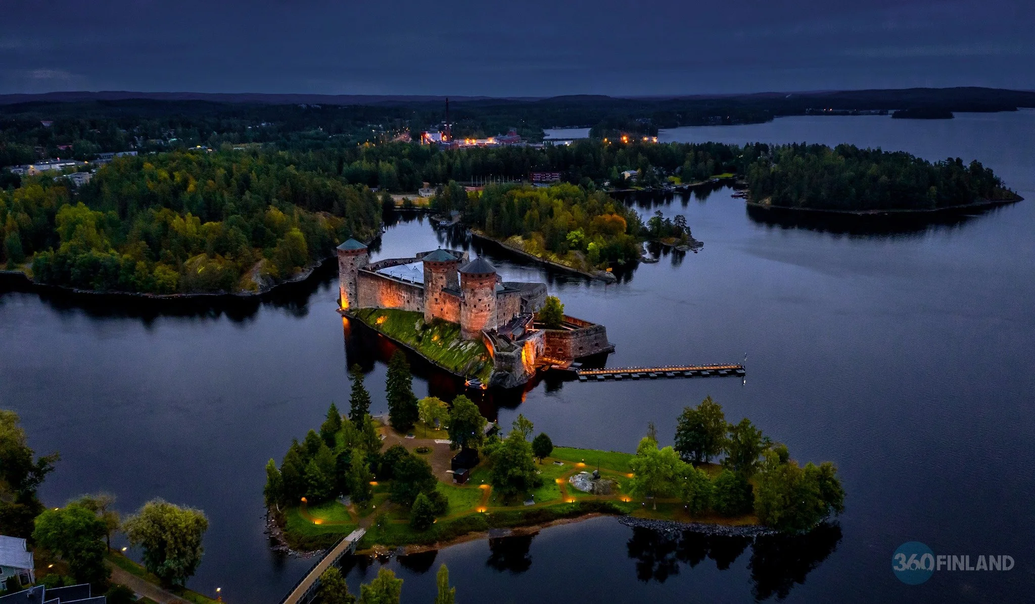 Aerial view of a historic castle on an island surrounded by water at dusk, with lights illuminating the structure and nearby walking paths.