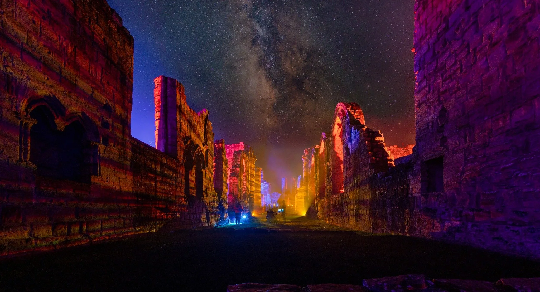 Ancient stone ruins illuminated with colorful lights at night under a starry sky with visible Milky Way.