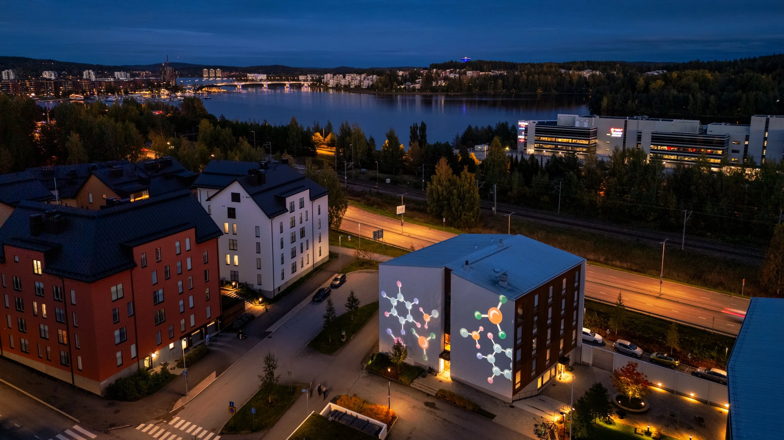 Aerial view of a cityscape at dusk with illuminated buildings, a river, and a street with cars. A residential building has digital molecular diagrams projected on its wall.