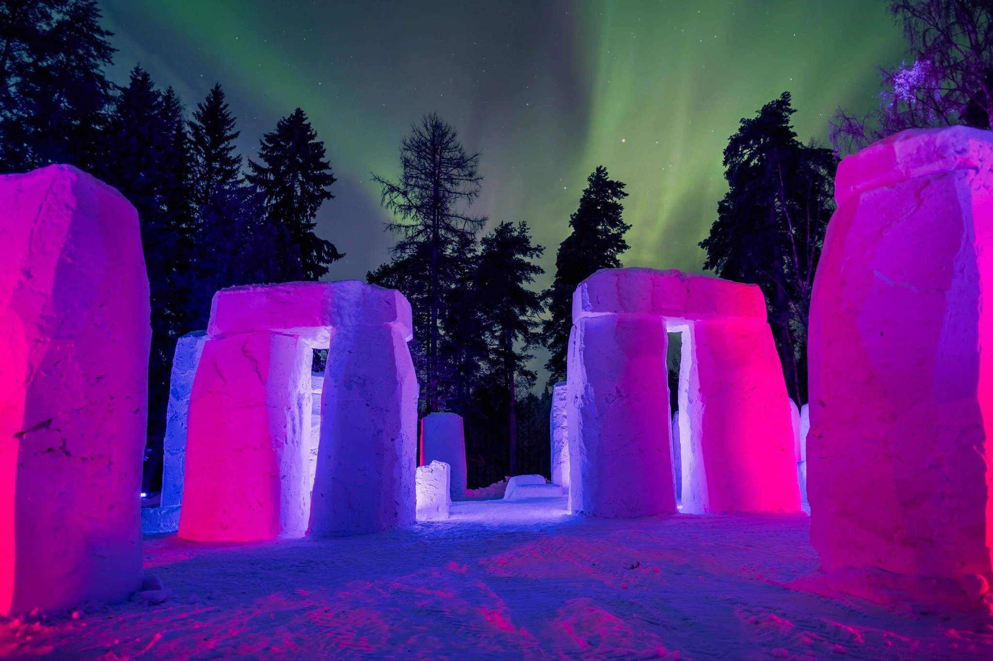 Ice sculptures illuminated with pink and purple lights set in a snowy landscape under a night sky with green aurora borealis and stars, surrounded by tall evergreen trees.