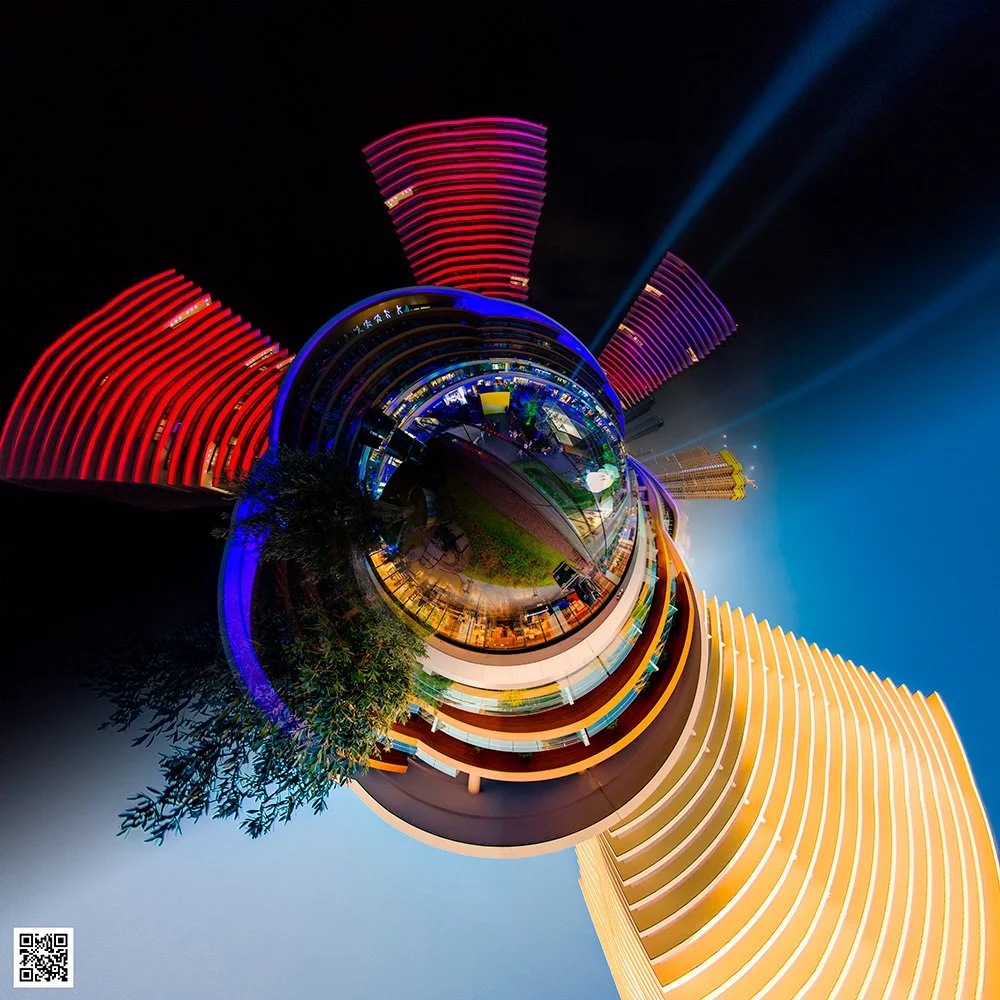 Spherical panoramic view of brightly lit modern buildings at night, featuring red and yellow lights, with a central area containing trees and pathways. The image has a stylized, planet-like appearance.