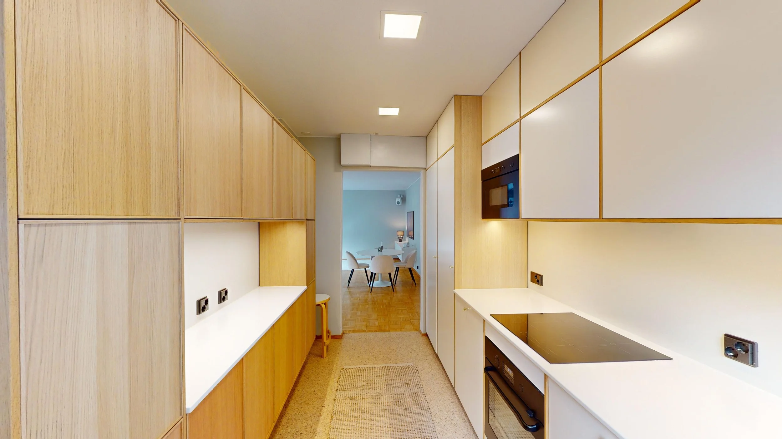 Modern kitchen with white and wood cabinets, black cooktop, and a view into a dining area with a round table and white chairs.