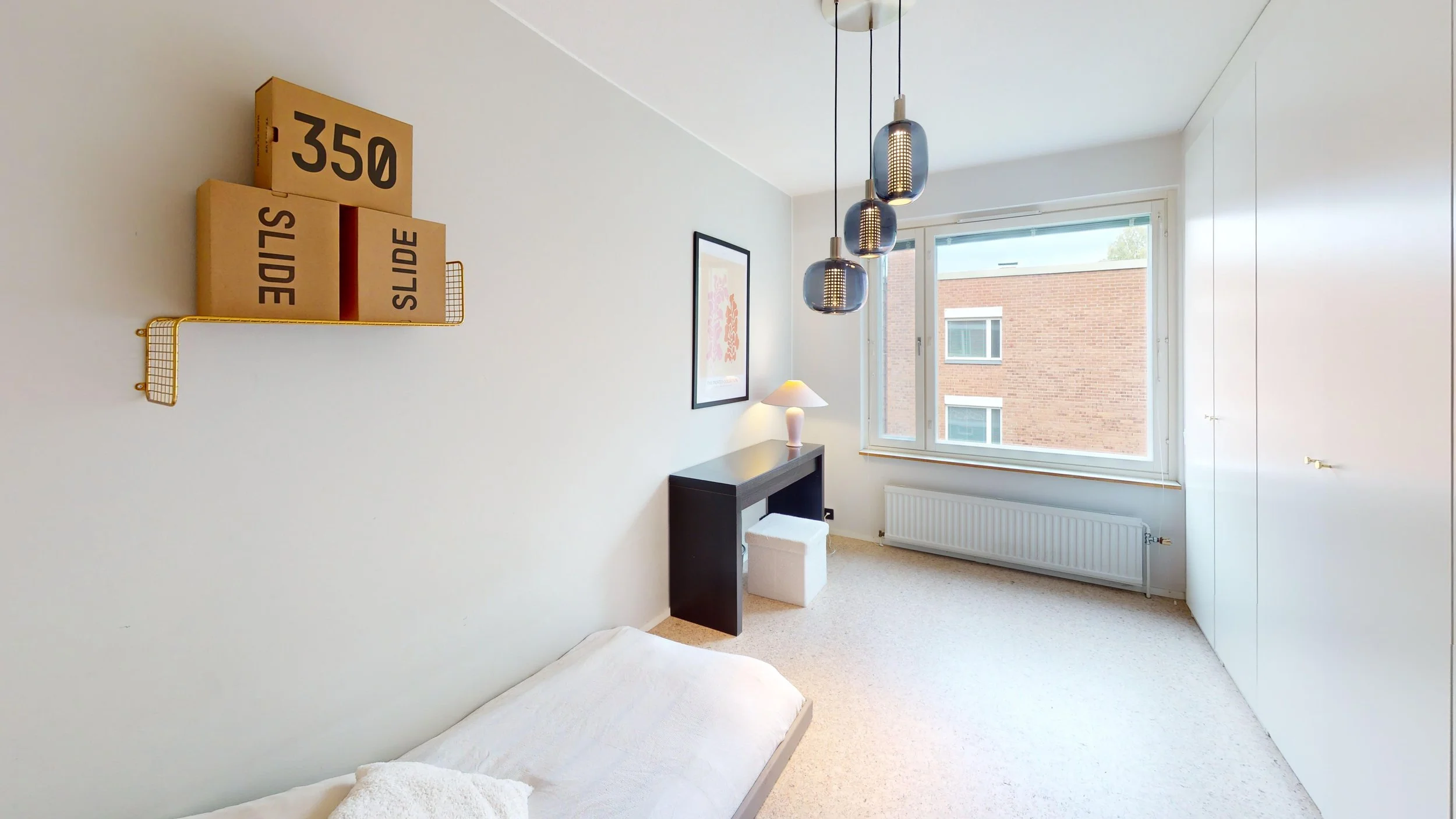 Minimalist bedroom with white walls, large window, black desk, white stool, orange lamp, framed art, black hanging pendant lights, and a wall shelf with cardboard boxes labeled '350' and 'SLIDE'.