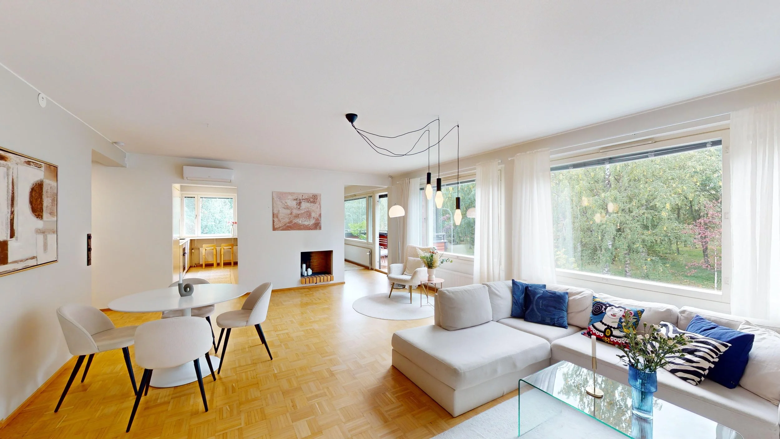 Bright living room with large windows, white sectional sofa with blue and patterned pillows, glass coffee table with blue vase and flowers, modern hanging pendant lights, and a round area rug. Visible dining area with white table and chairs, small no