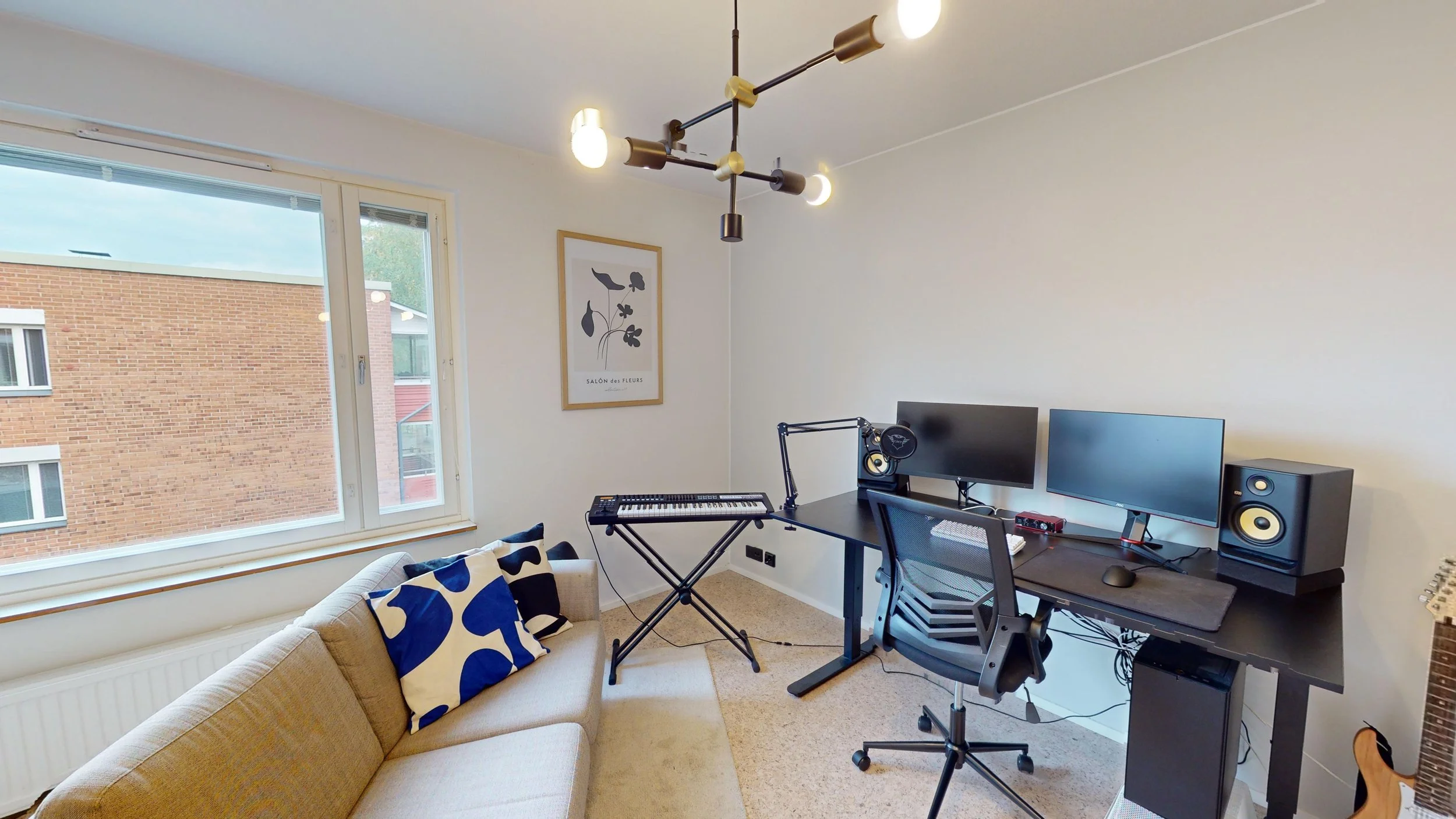 A modern home office with a beige sofa, blue and white pillows, a large window, and a black desk with dual monitors, a keyboard, and a speaker. There is a keyboard on a stand, a microphone, a chair, and a geometric chandelier hanging from the ceiling