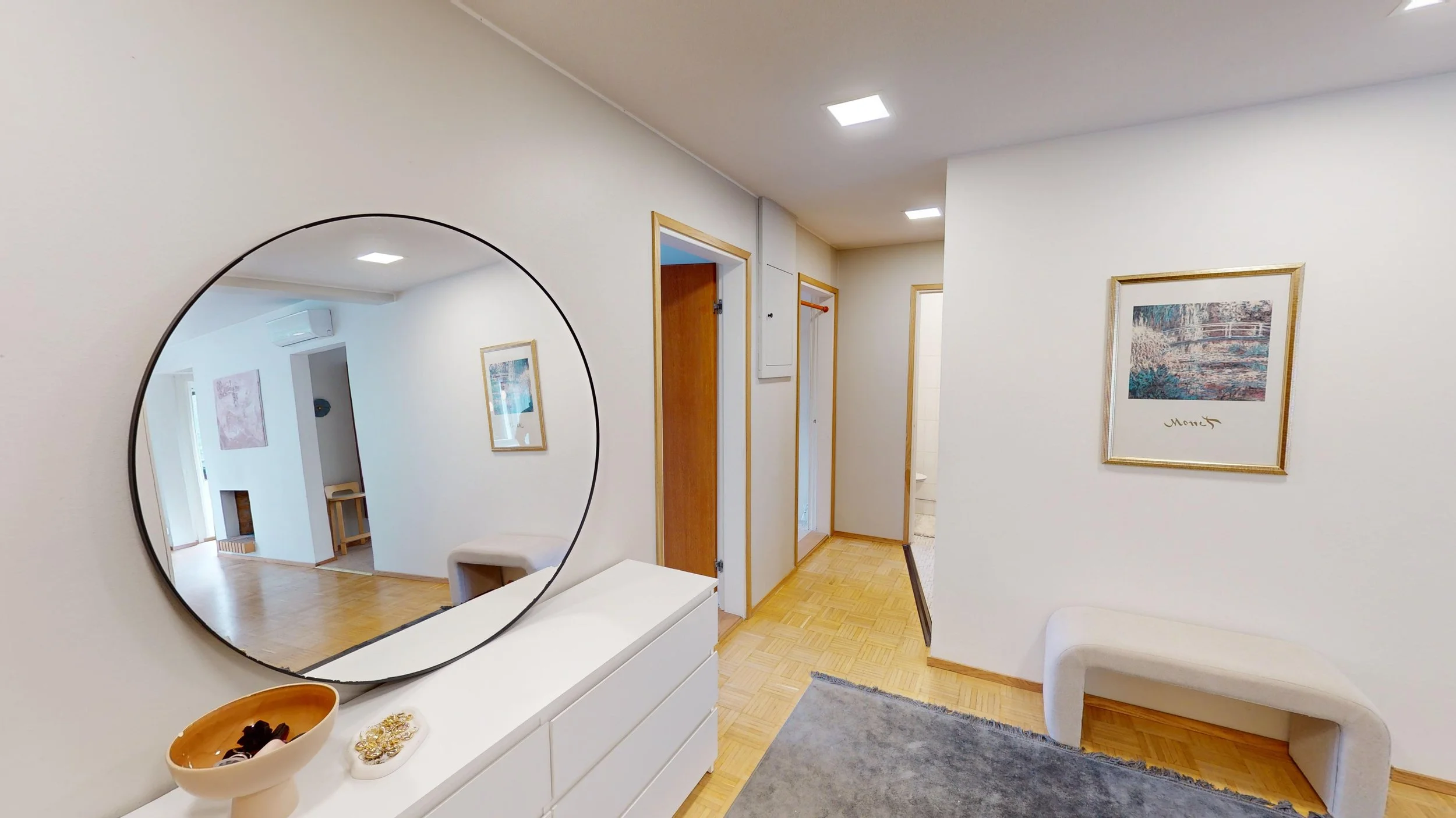 A hallway with white walls and wooden floors, a large round mirror on the left wall, artwork on the walls, a white dresser underneath the mirror, and a curved white bench on the right, leading to other rooms.