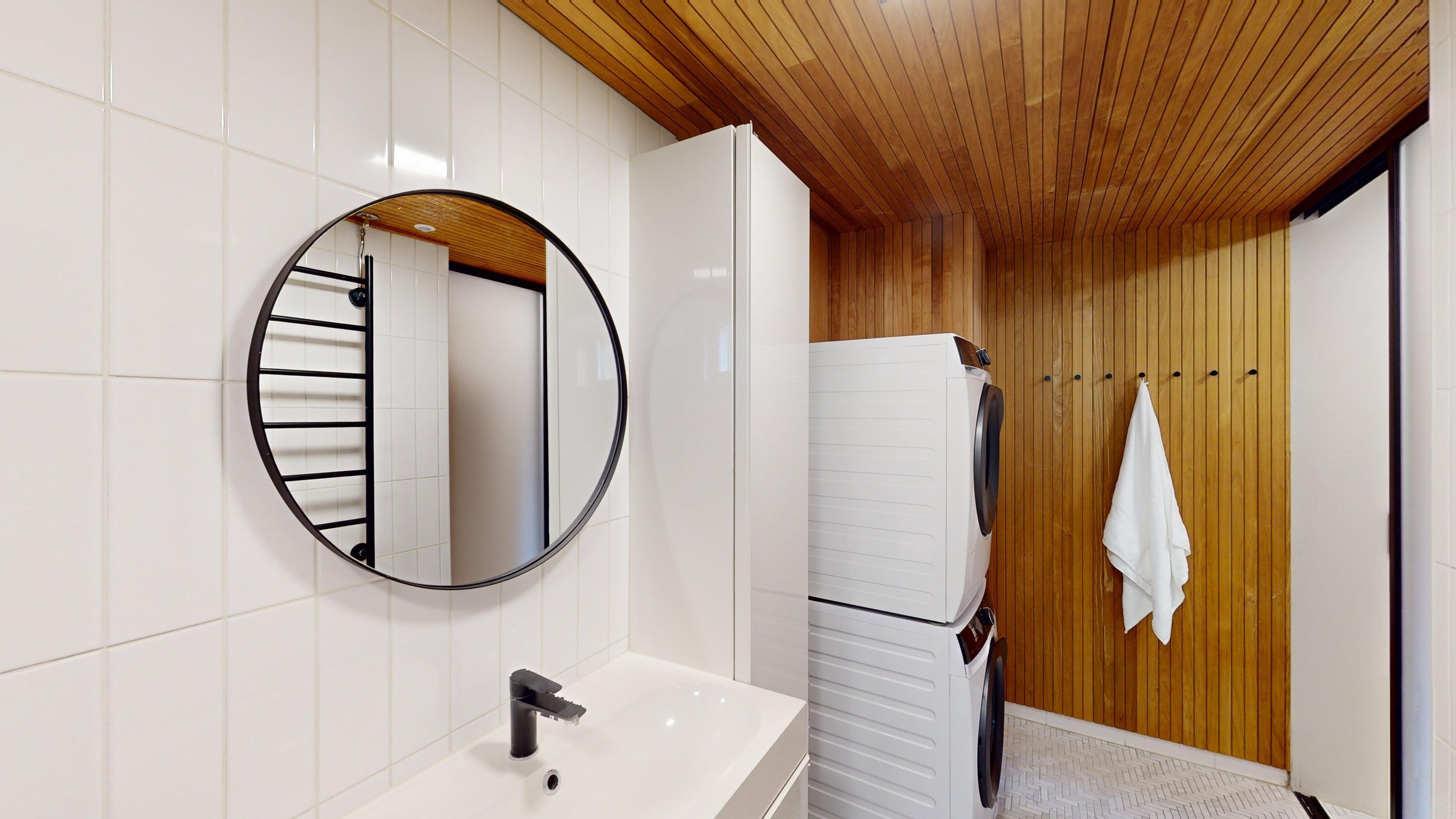 A laundry room with a white washing machine and dryer stacked against a wooden wall. A white towel hangs on a hook on the wooden wall. There is a white sink with a black faucet, and a round mirror above the sink reflecting part of the laundry room. T