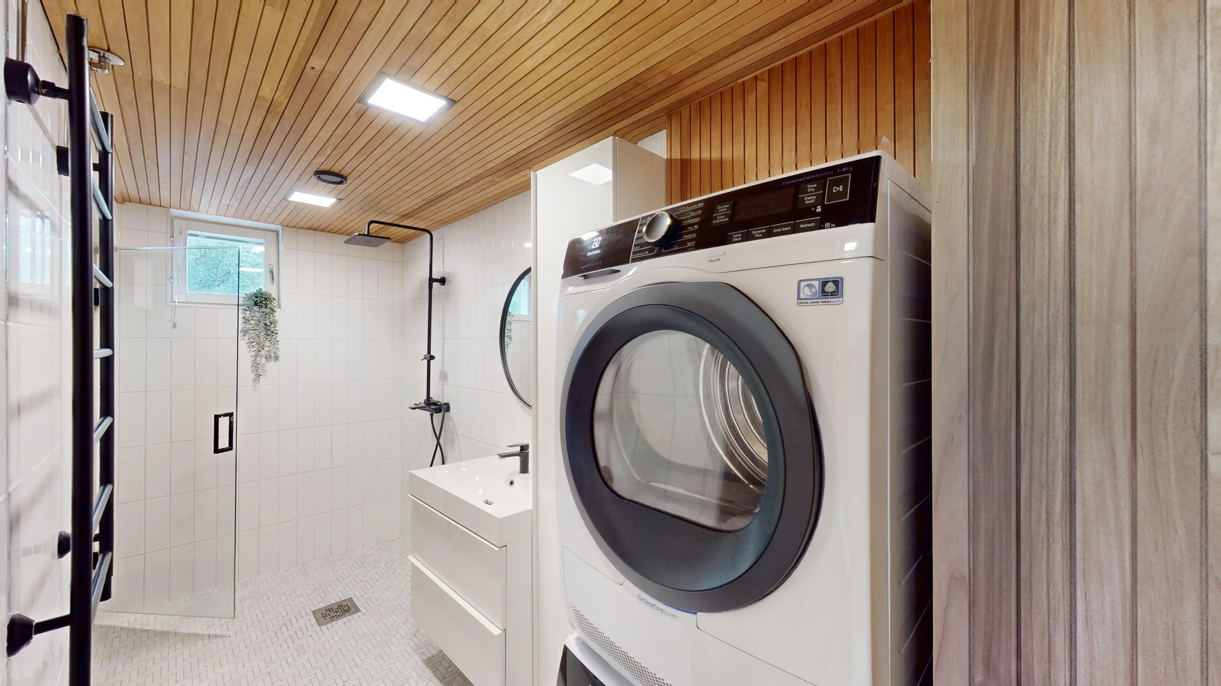 Modern laundry room with front-loading washing machine, small sink, walk-in shower, and wooden ceiling.