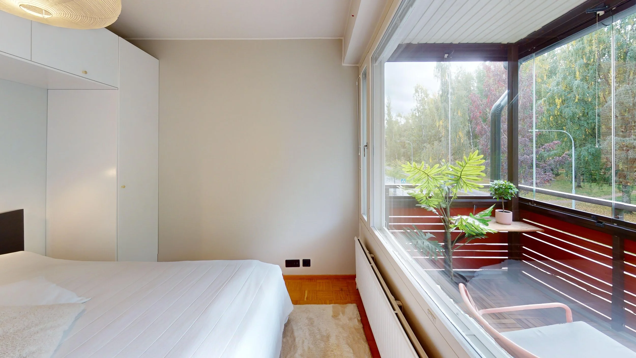 A bedroom with white walls and a large window opening onto a balcony with potted plants, a wooden deck, and trees outside.
