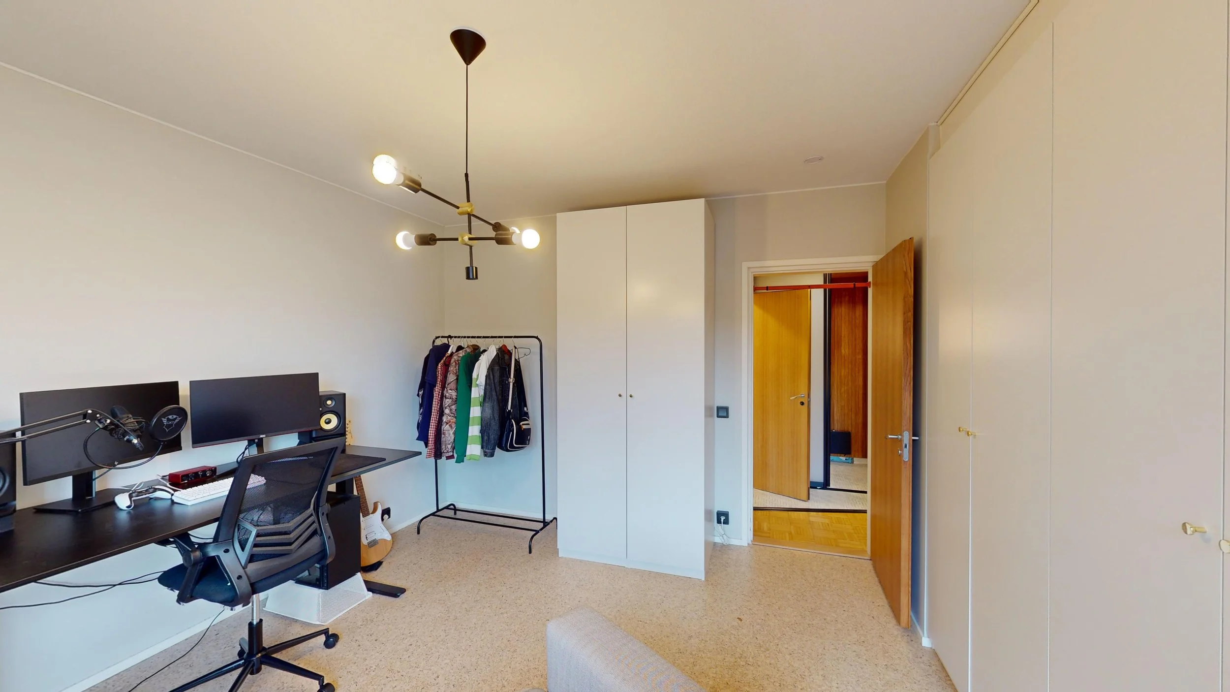 Home office with dual monitors, microphone, and speaker, a clothing rack with clothes, and wooden doors leading to another room.