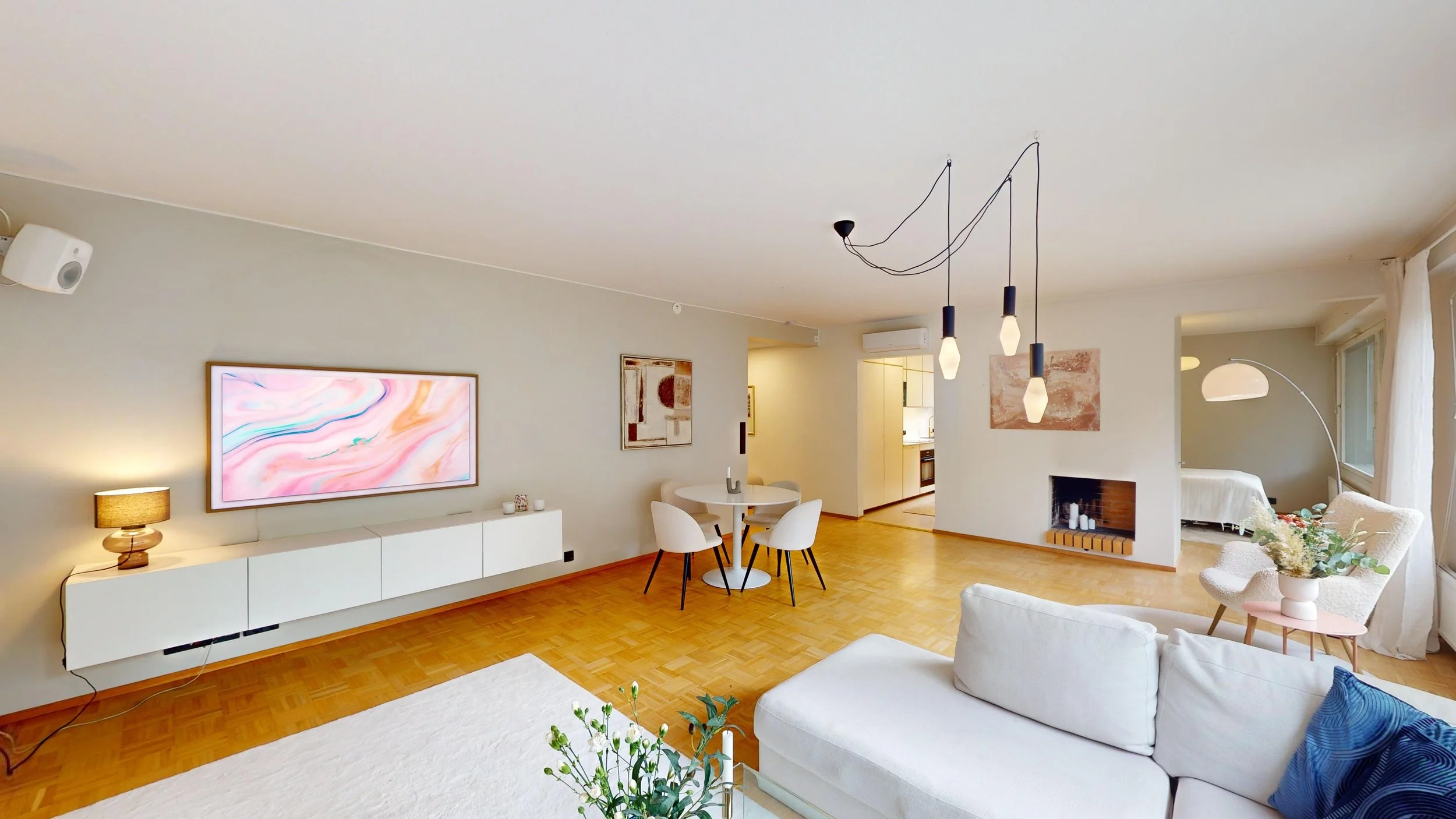 Bright living room with white sofa, dining area with white table and chairs, a wall-mounted TV, a brick fireplace, and a kitchen in the background. Large windows with curtains, modern hanging lights, and decorative items.