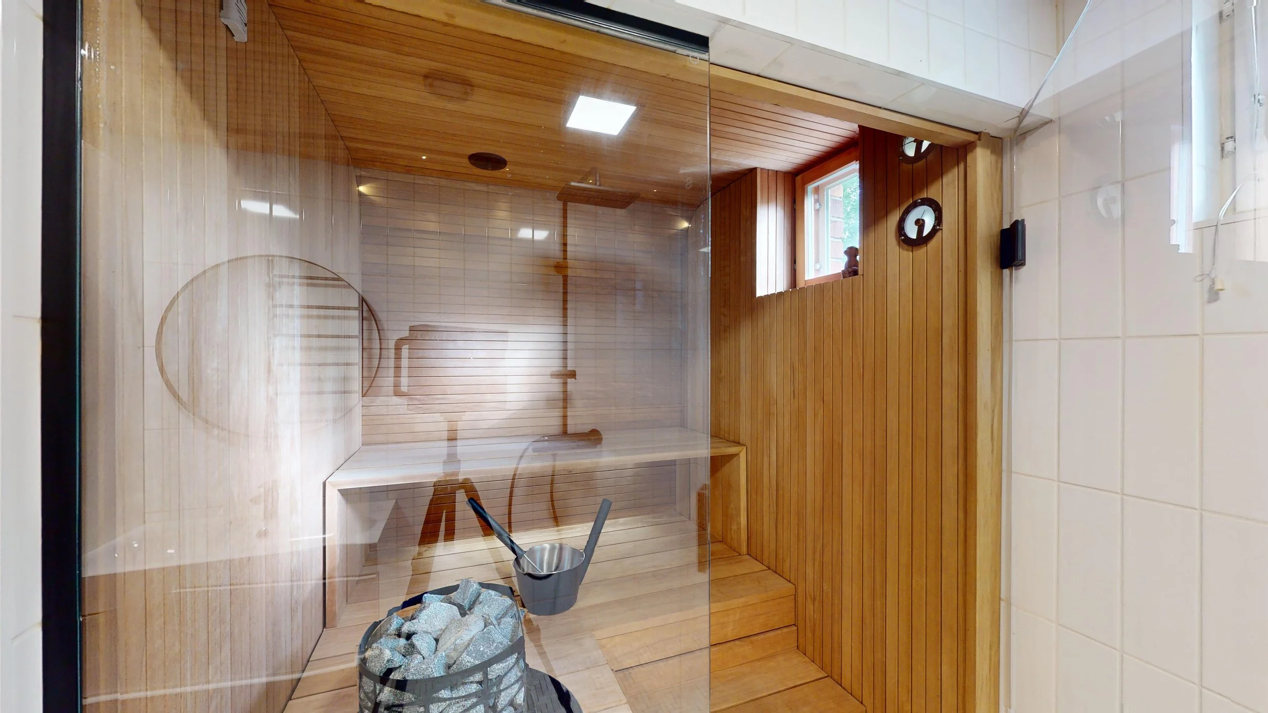 A sauna room with wooden walls and floor, a small window, a wall clock, a bucket with a ladle, and a stone heater with water.