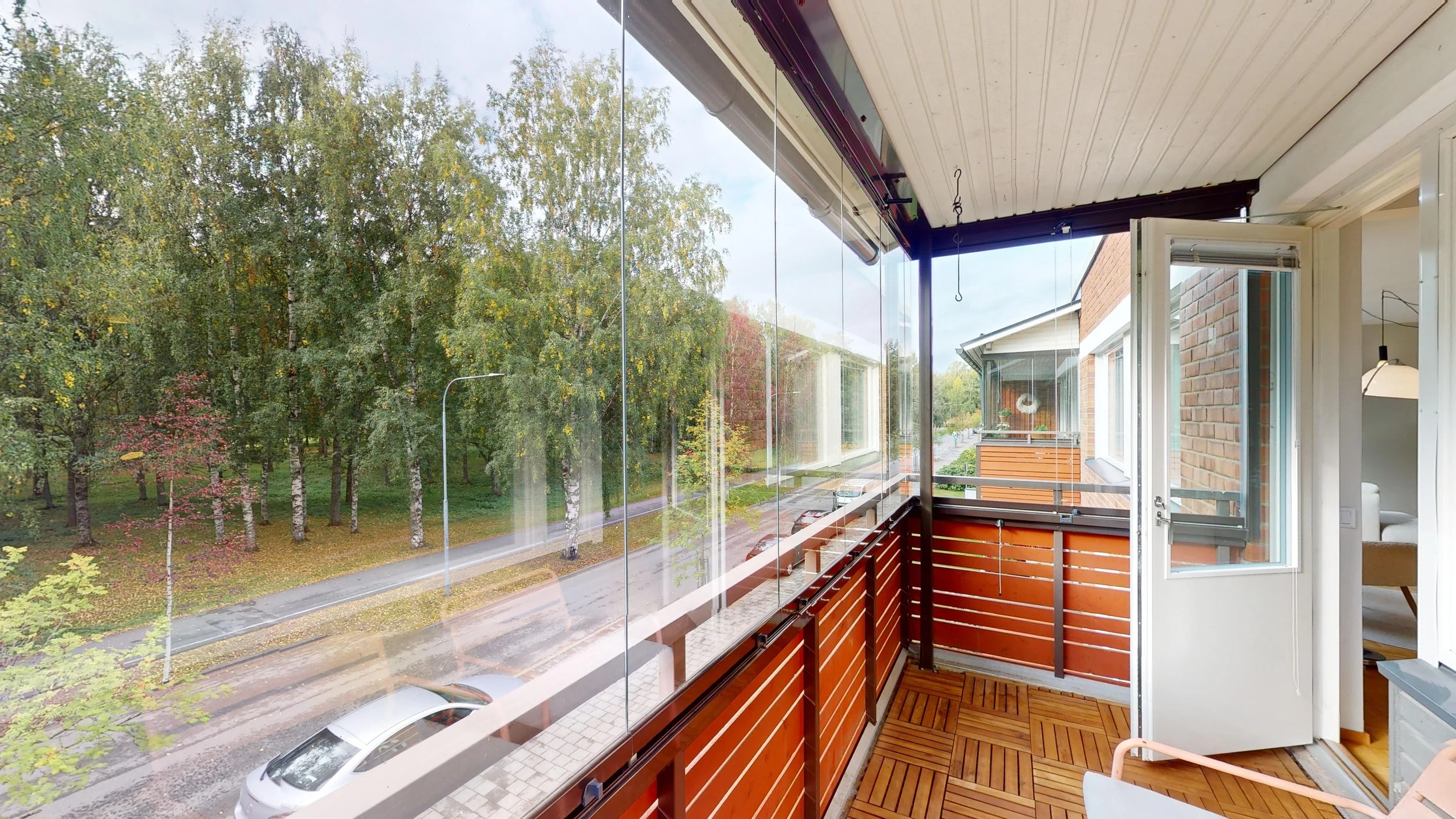 View from a balcony overlooking a street lined with trees, with a parked silver car visible below. The balcony has wooden flooring and a railing with horizontal slats, and an open door leads inside a room with a modern lamp and furniture visible.