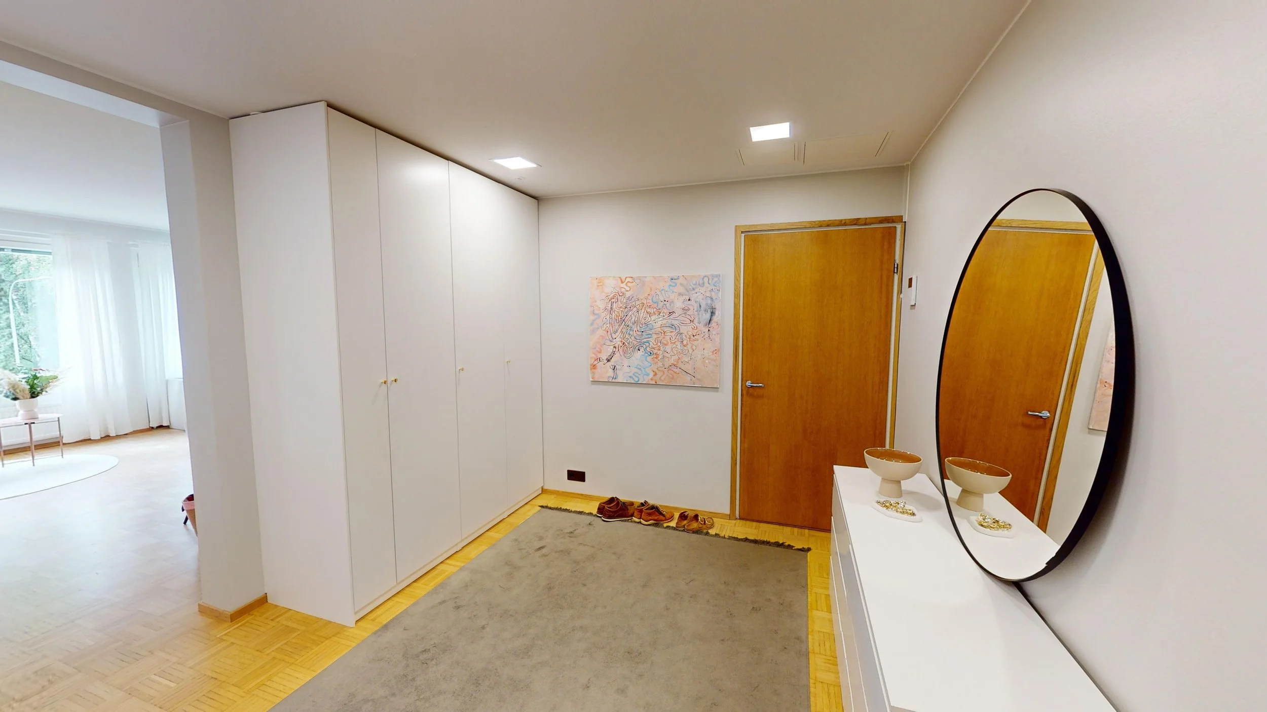 Entryway with wooden door, white cabinets, large mirror, rugs, shoes, and abstract wall art in a modern home.