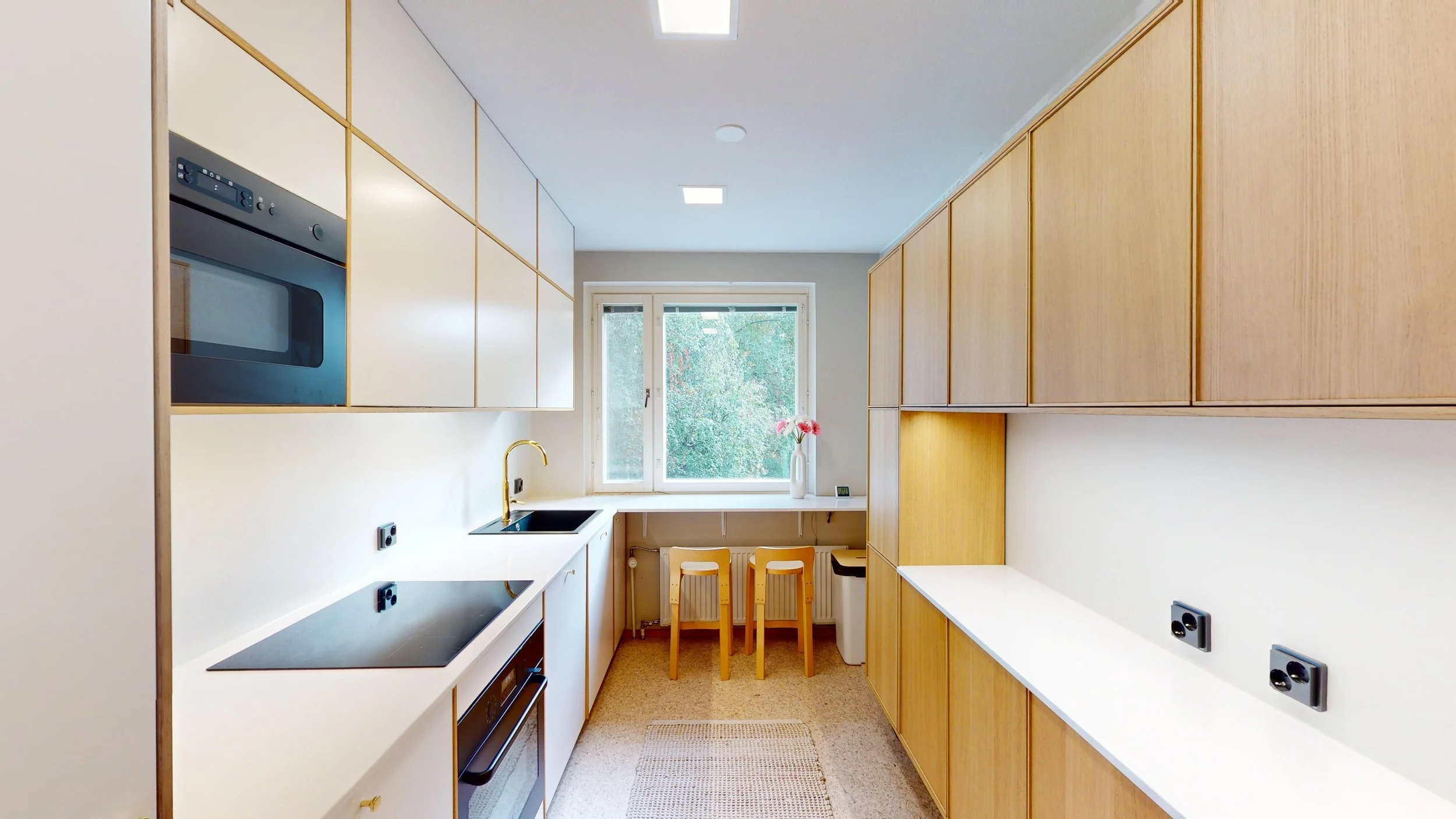 A modern kitchen with wooden cabinets, white countertops, a black electric stovetop, a built-in microwave, a gold faucet, a sink, a window with two chairs and a vase with pink flowers, and beige carpeted floor.