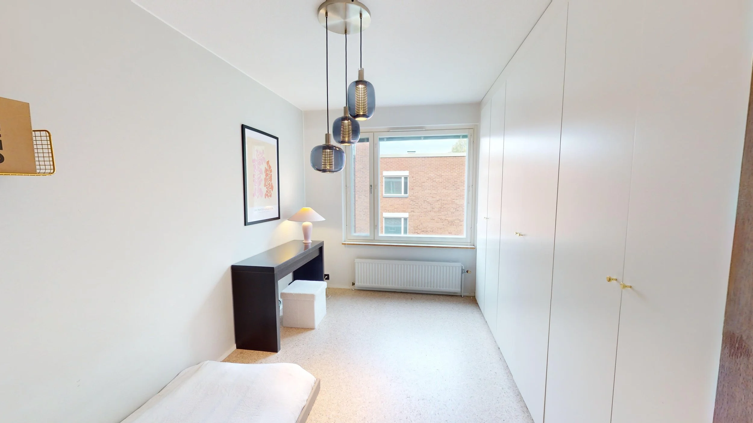 A minimalist bedroom with a large window, white walls, a black desk with a white lamp, a framed artwork, a white bed, and built-in white cabinets with small gold knobs.