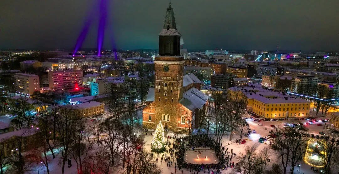 Aerial view of a snowy city Turku at night featuring a large cathedral with a lit Christmas tree in front, surrounded by people and colorful lights.