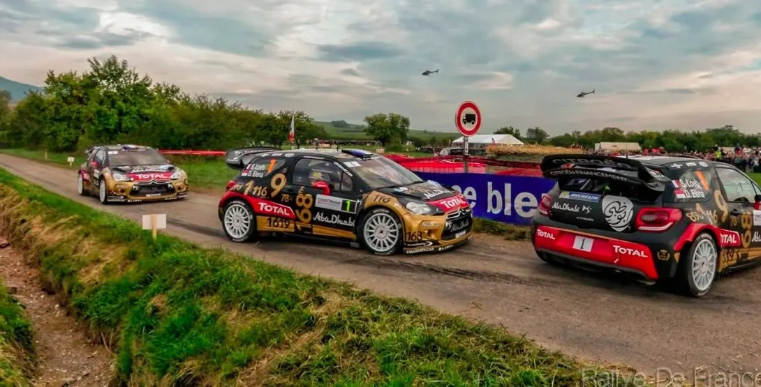 Rally cars with Total and Abu Dhabi sponsorship on a rural road during a motorsport event, with spectators and helicopters in the background.