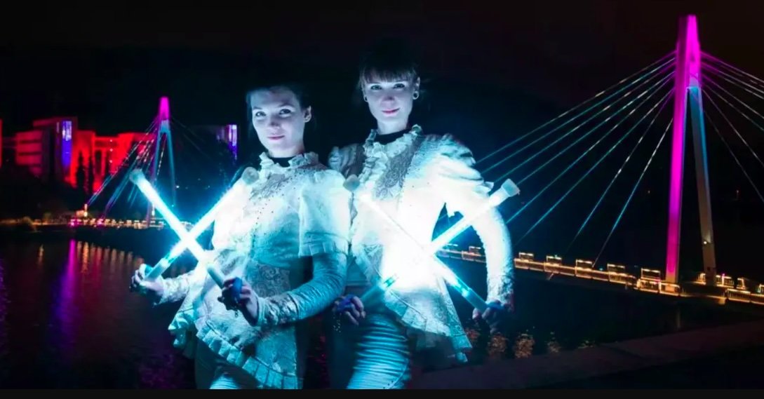 Two people in white outfits holding illuminated sticks at night with a lit-up cable-stayed bridge in the background.