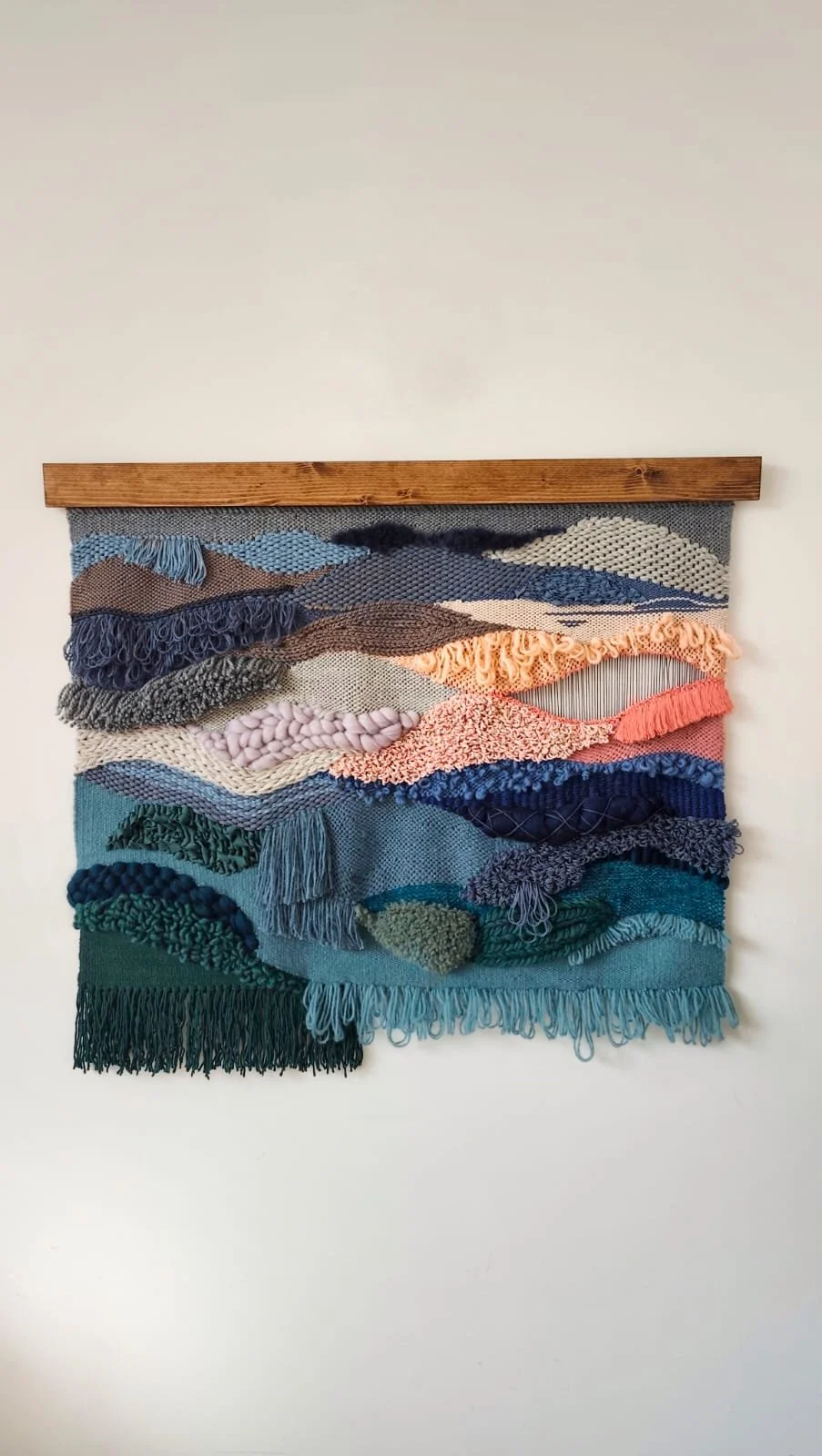 Colorful textile wall hanging with layered yarn and fabric waves, mounted on a wooden rod, against a white wall. Weaving technique.