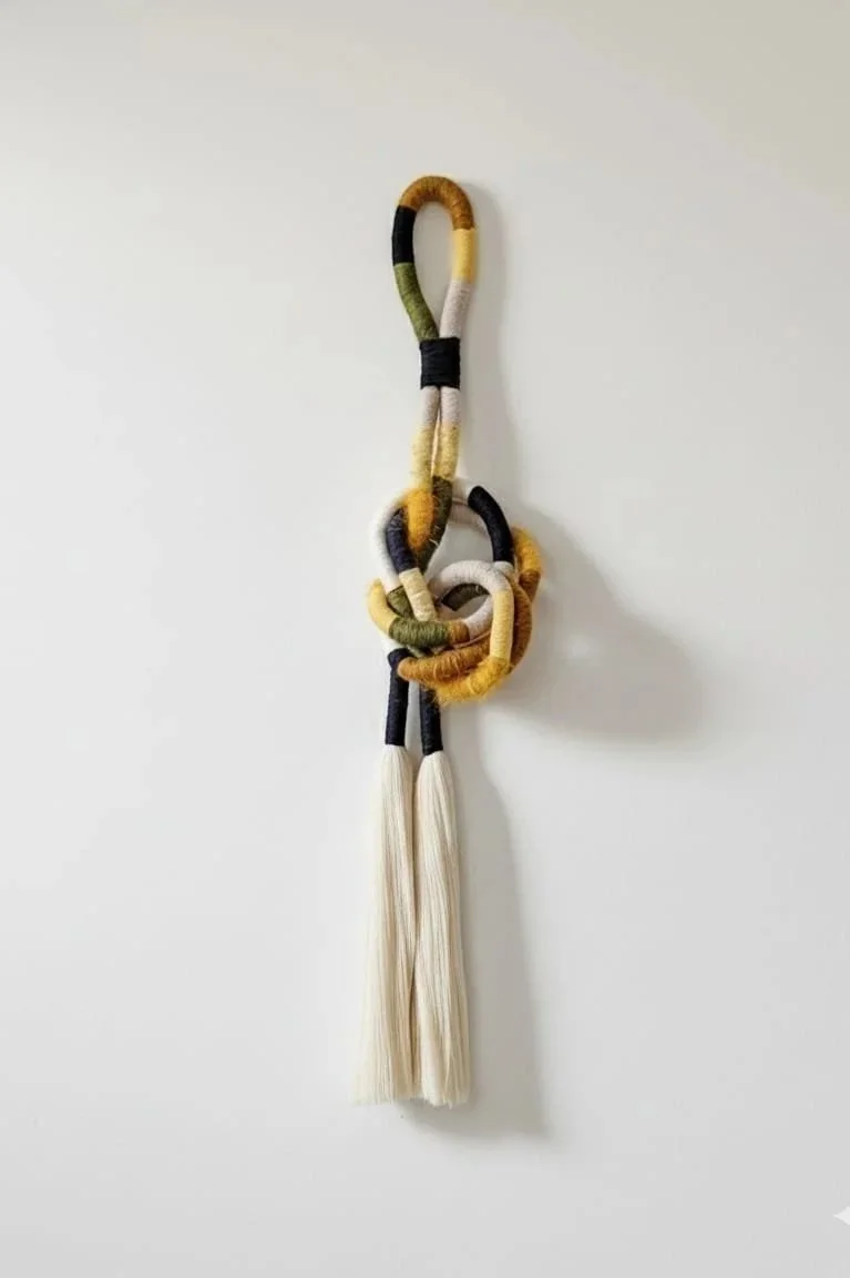 A decorative wall hanging made of intertwined and looped colorful yarn with tassels at the bottom, secured with a black band near the top.