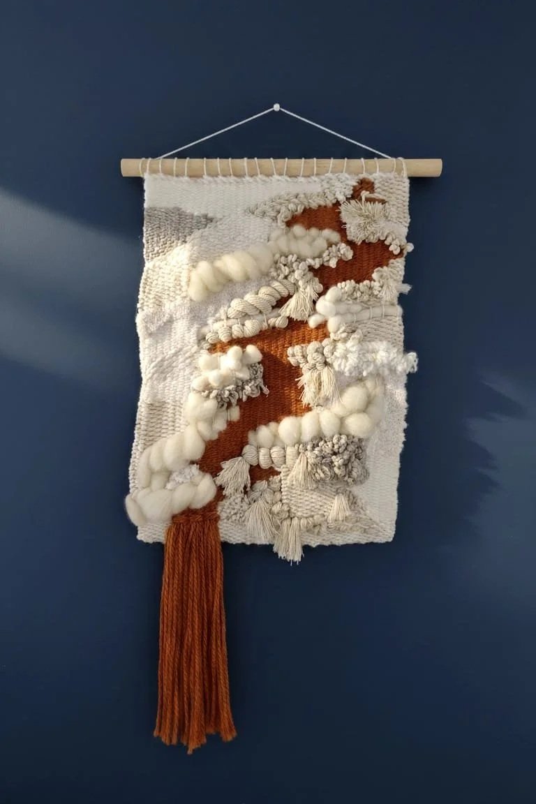 Textured wall hanging with yarn and fabric in white, cream, and rust colors, suspended on a wooden rod against a dark blue wall.