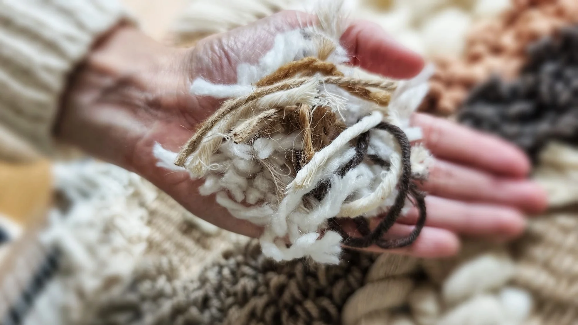 Close-up of a hand holding a bundle of mixed textile fibers, including yarn and fabric scraps, with other similar bundles in the background.