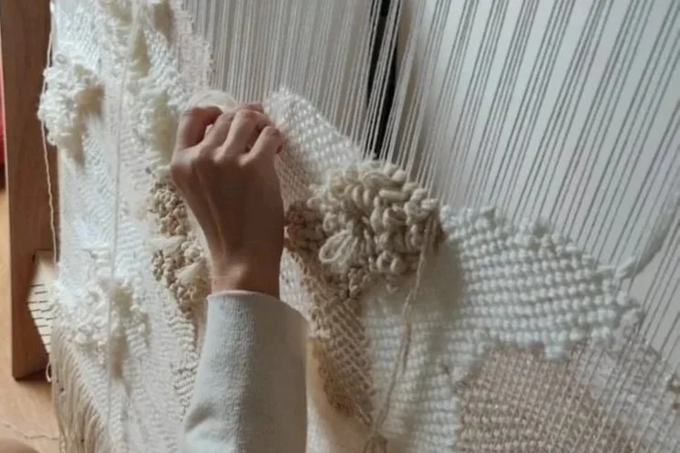 Person weaving or creating a textile wall hanging on a loom with cream-colored yarn.