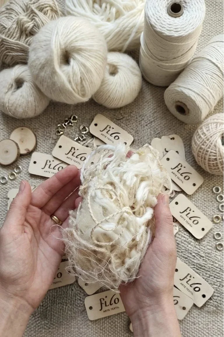 Hands holding a tangled ball of cream-colored yarn surrounded by skeins of yarn, small tags with the brand name 'FILO,' and small decorative buttons and metal rings on a textured fabric surface.