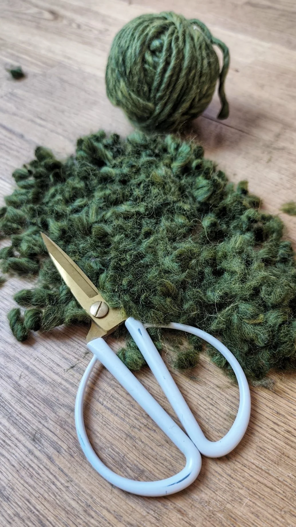A ball of green yarn, a pair of white-handled scissors, and a pile of green yarn scraps on a wooden surface.