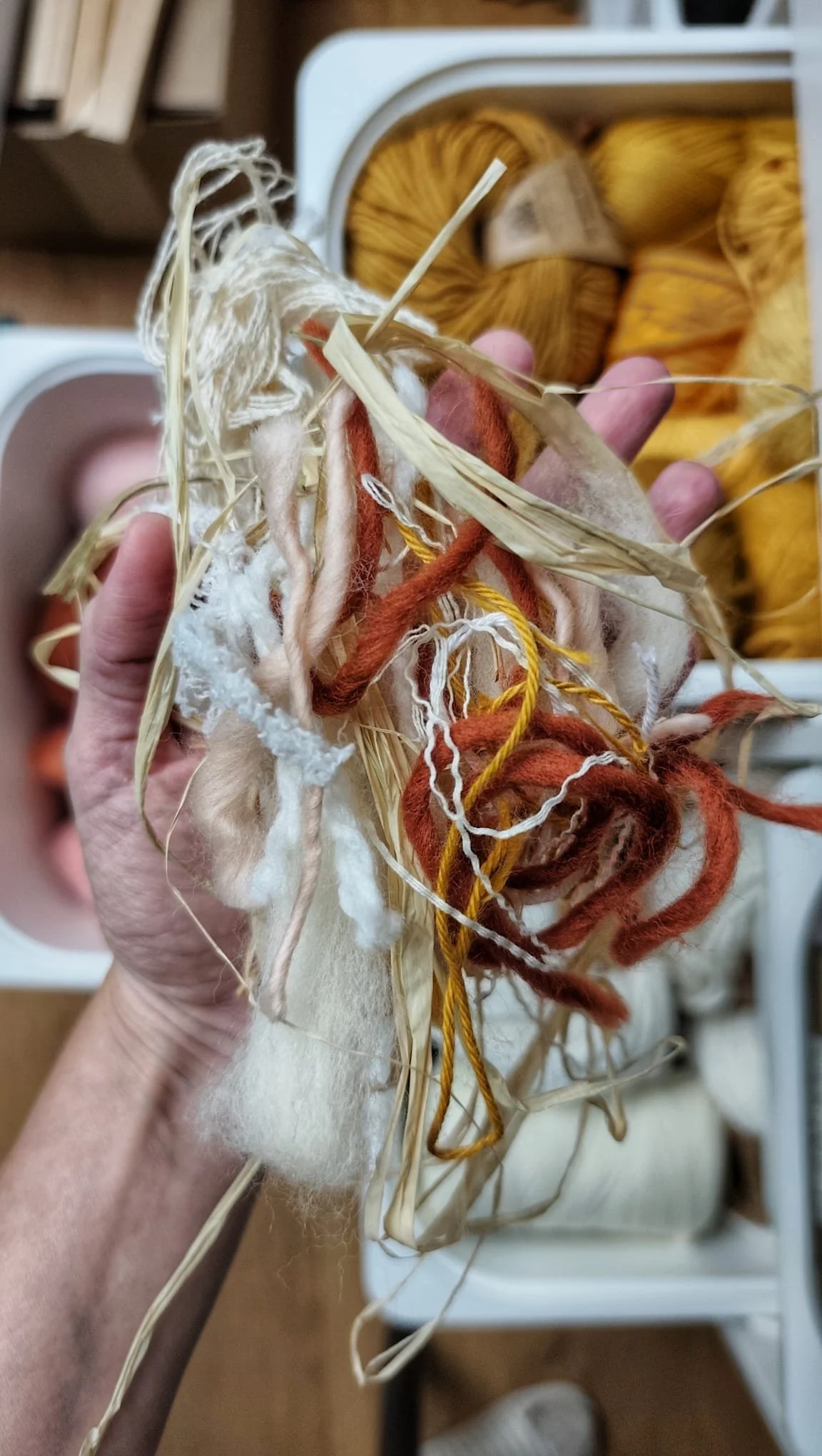 Hand holding various colored yarns and fiber amongst straw in front of a box of yellow yarn balls.