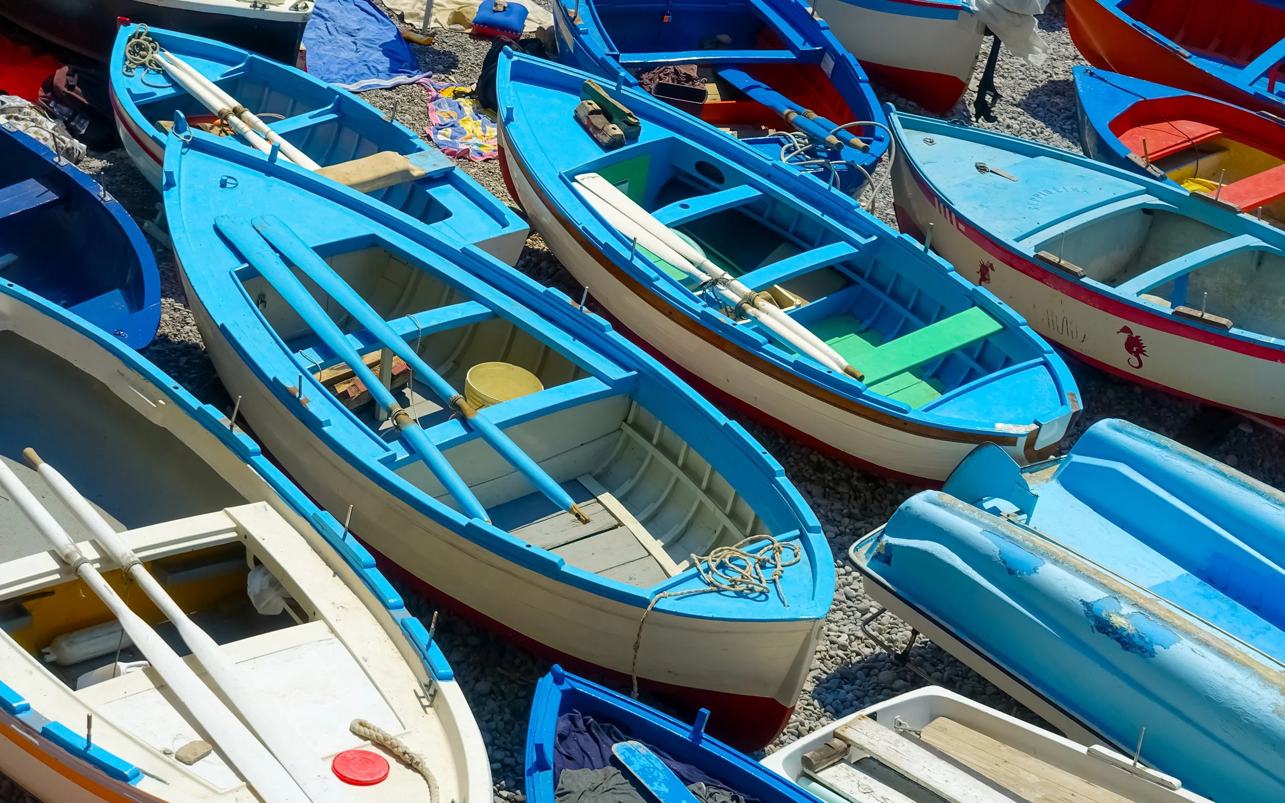 Several small rowboats in various colors, primarily blue, stored on a rocky beach, with some equipment and personal items inside and around the boats.