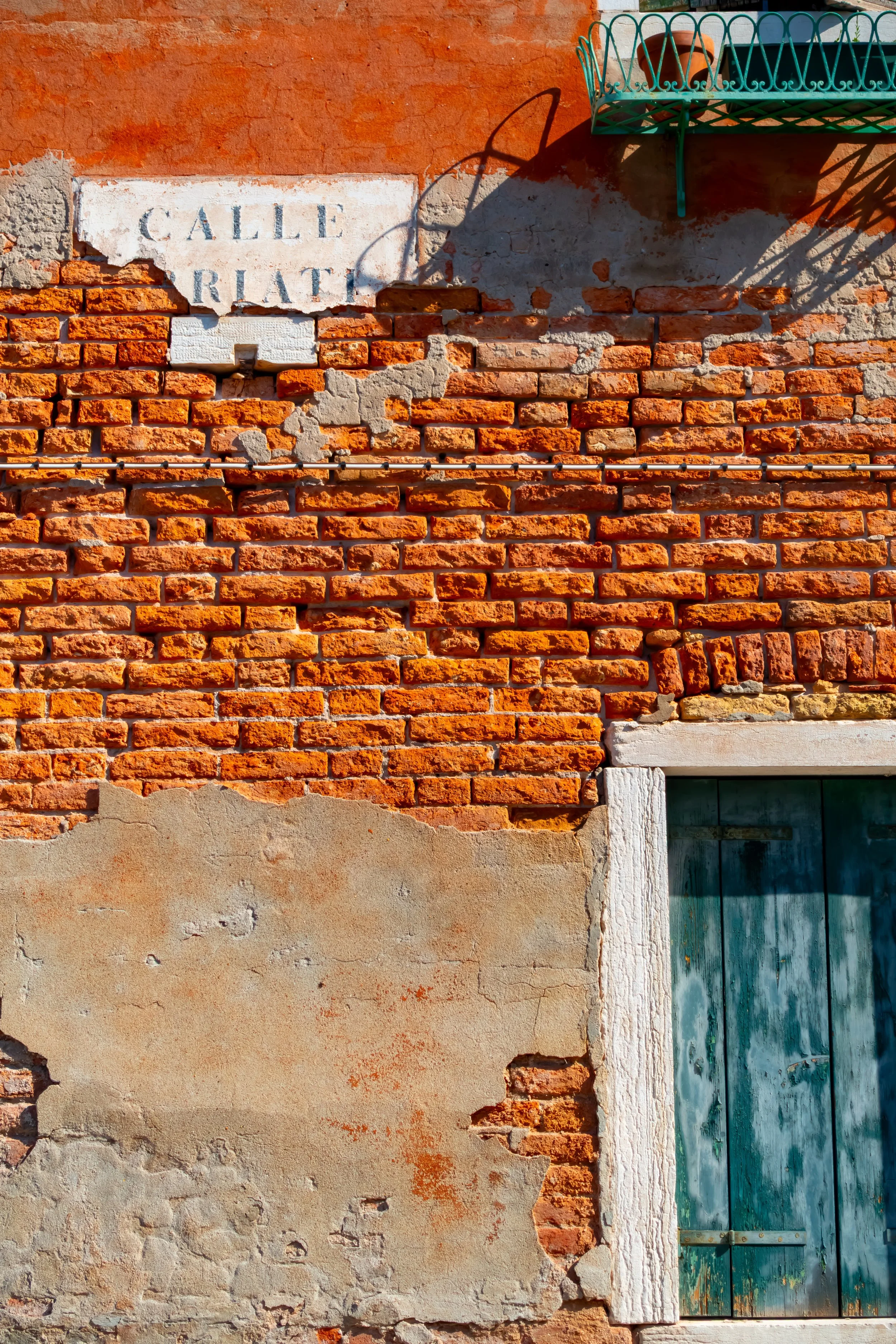 A weathered brick wall with a patch of peeling plaster and a green wooden door. A street sign reading 'Calle Rialto' is mounted on the wall. There is a small green metal balcony with a decorative railing, casting a shadow.