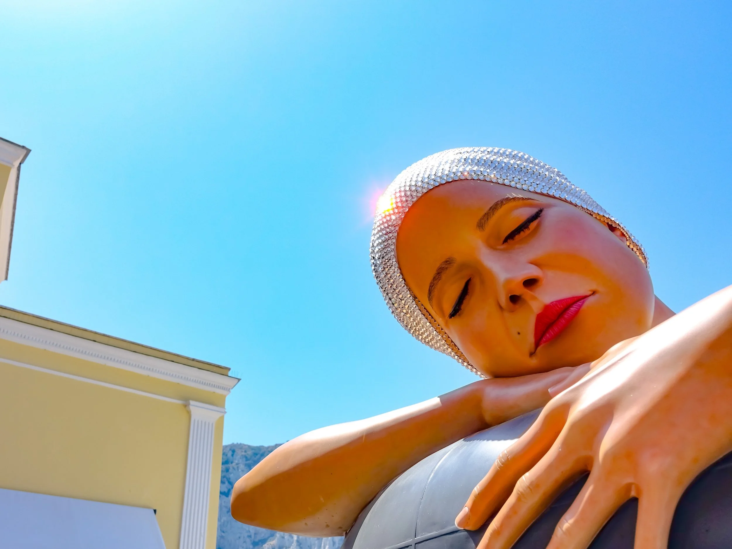 Close-up of a woman with a sparkling headscarf, eyes closed, resting her chin on her crossed arms, against a bright blue sky with a yellow building and a mountain in the background.