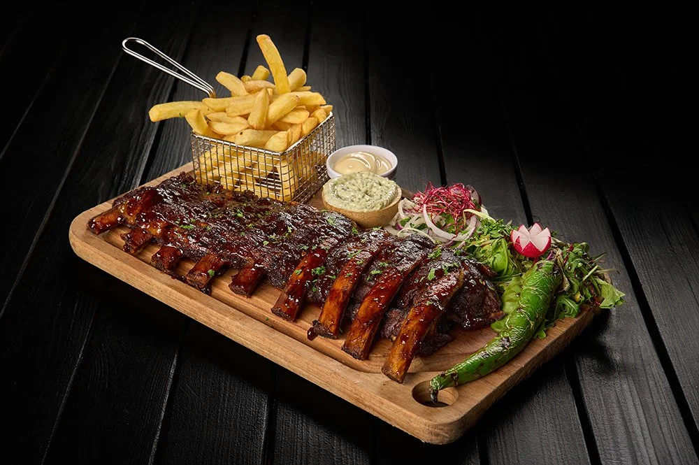 Varkensribben met barbecuesaus, gepofte aardappels met kruidenboter, gemengde salade, en frietjes op een houten snijplank, met een donkere achtergrond.