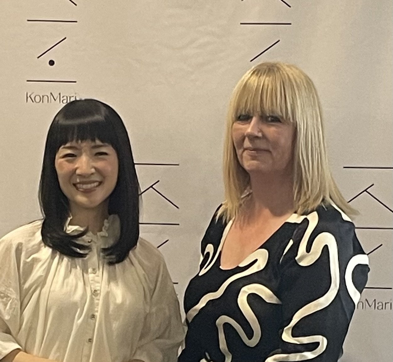 Two women standing together in front of a beige background with a partial clock and text, smiling at the camera.