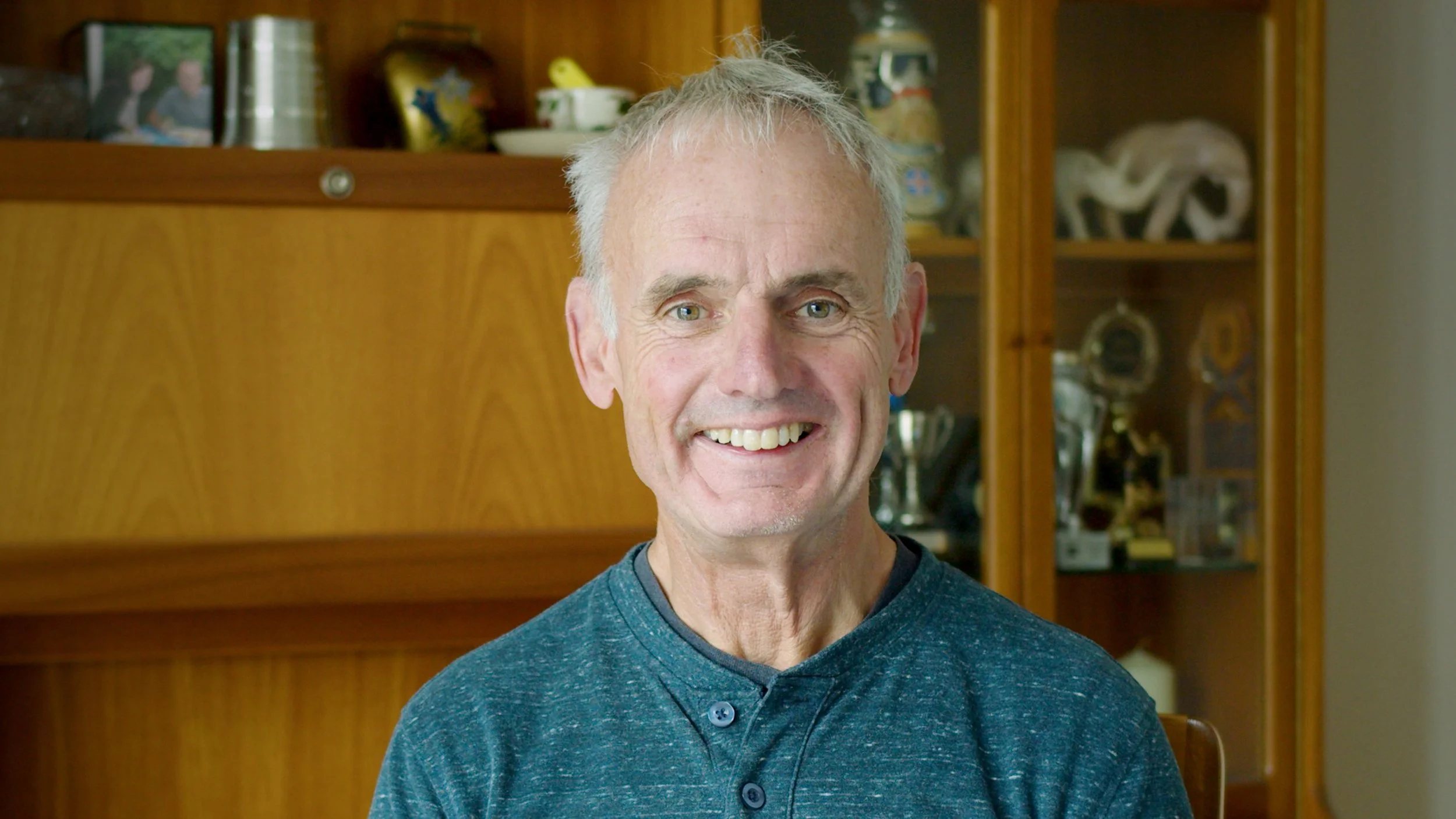 A smiling middle-aged man with gray hair and blue eyes, wearing a blue button-up shirt, standing in front of wooden shelves filled with decorative items and trophies.