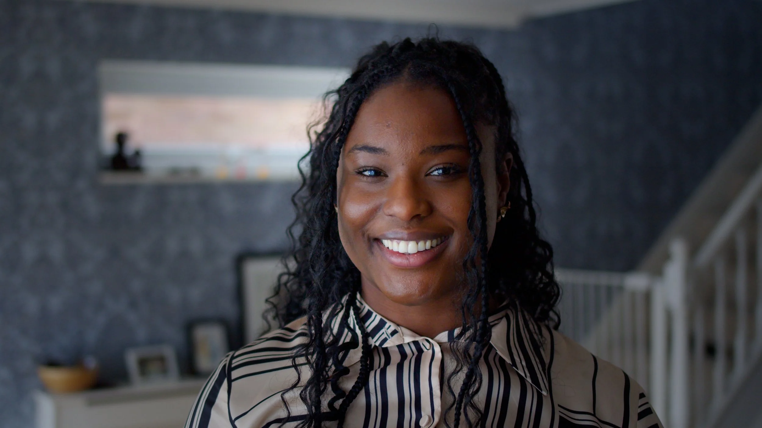 A woman with curly black hair smiling indoors with a kitchen background.