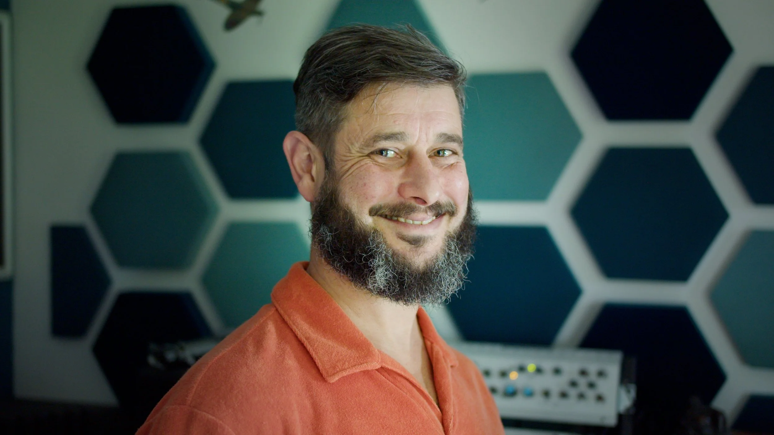 A smiling man with a beard and mustache, wearing an orange shirt, stands in front of a wall with a pattern of black hexagons on a white background, with audio mixing equipment in the background.