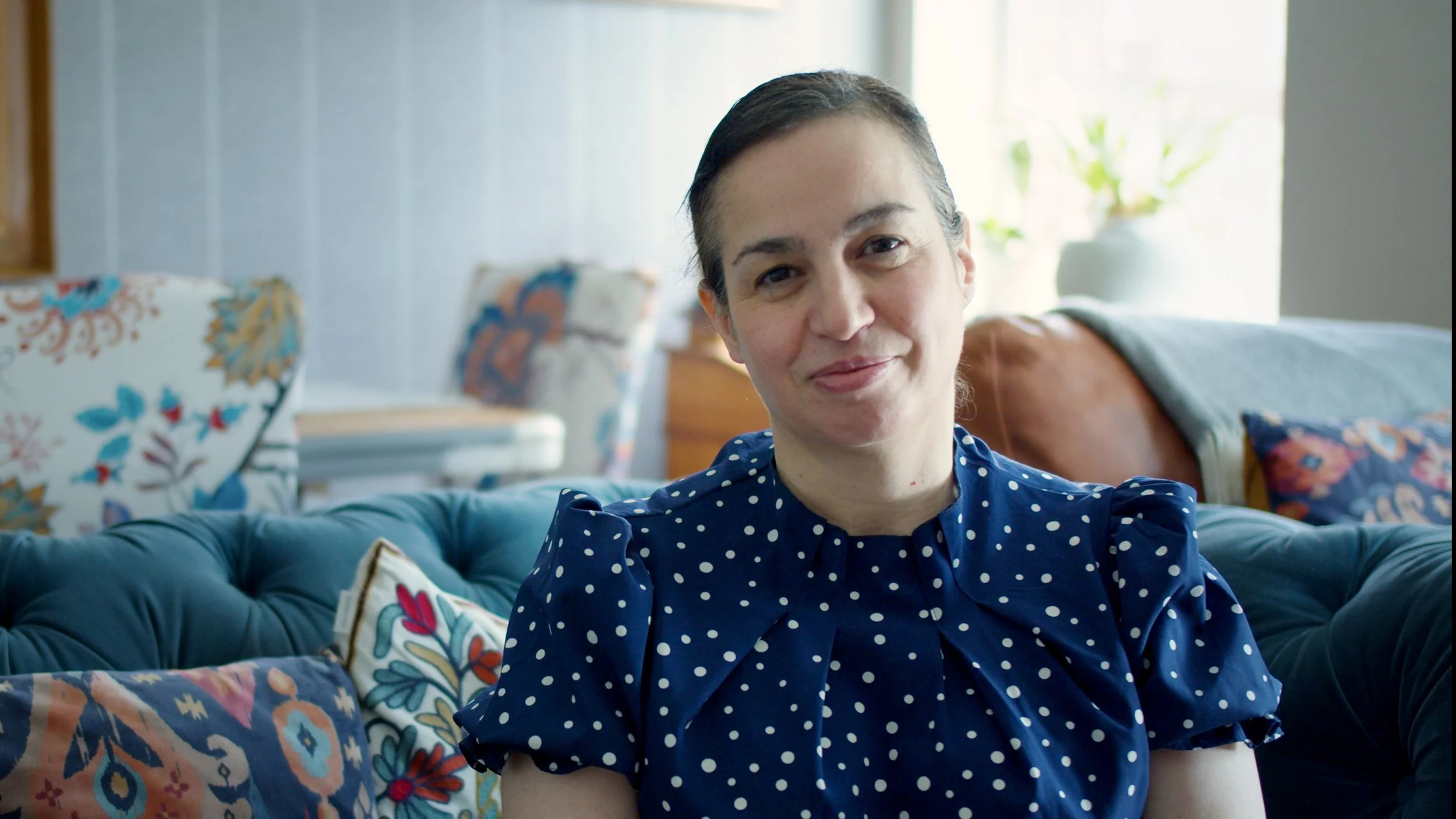 A woman with dark hair pulled back, wearing a navy blue polka dot blouse, sitting on a blue tufted sofa in a bright living room, smiling at the camera. There are colorful pillows on the sofa and a plant in the background near a window.